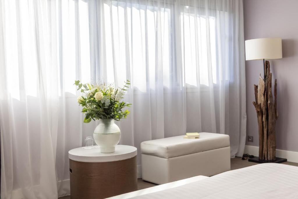 A hotel room with a bed, a round wooden side table with a vase of flowers, a white ottoman, sheer curtains, and a tall floor lamp.