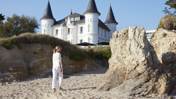 Un edificio parecido a un castillo con varias torres, una playa de arena y un acantilado rocoso.