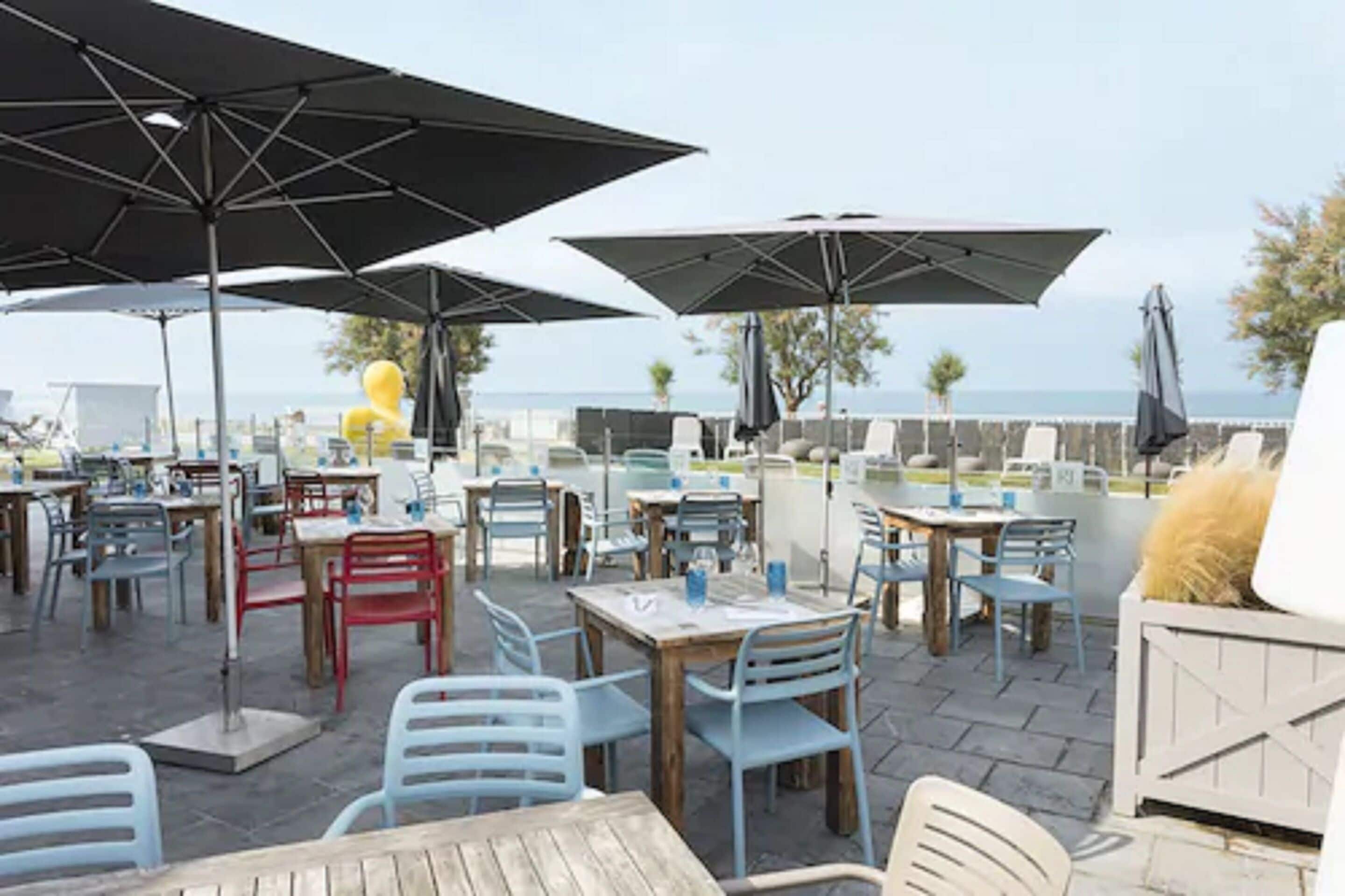 An outdoor dining area with tables and chairs, umbrellas, and a view of the sea.