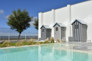 Zona de piscina con vistas despejadas al mar, un edificio blanco con detalles grises y un árbol en primer plano.