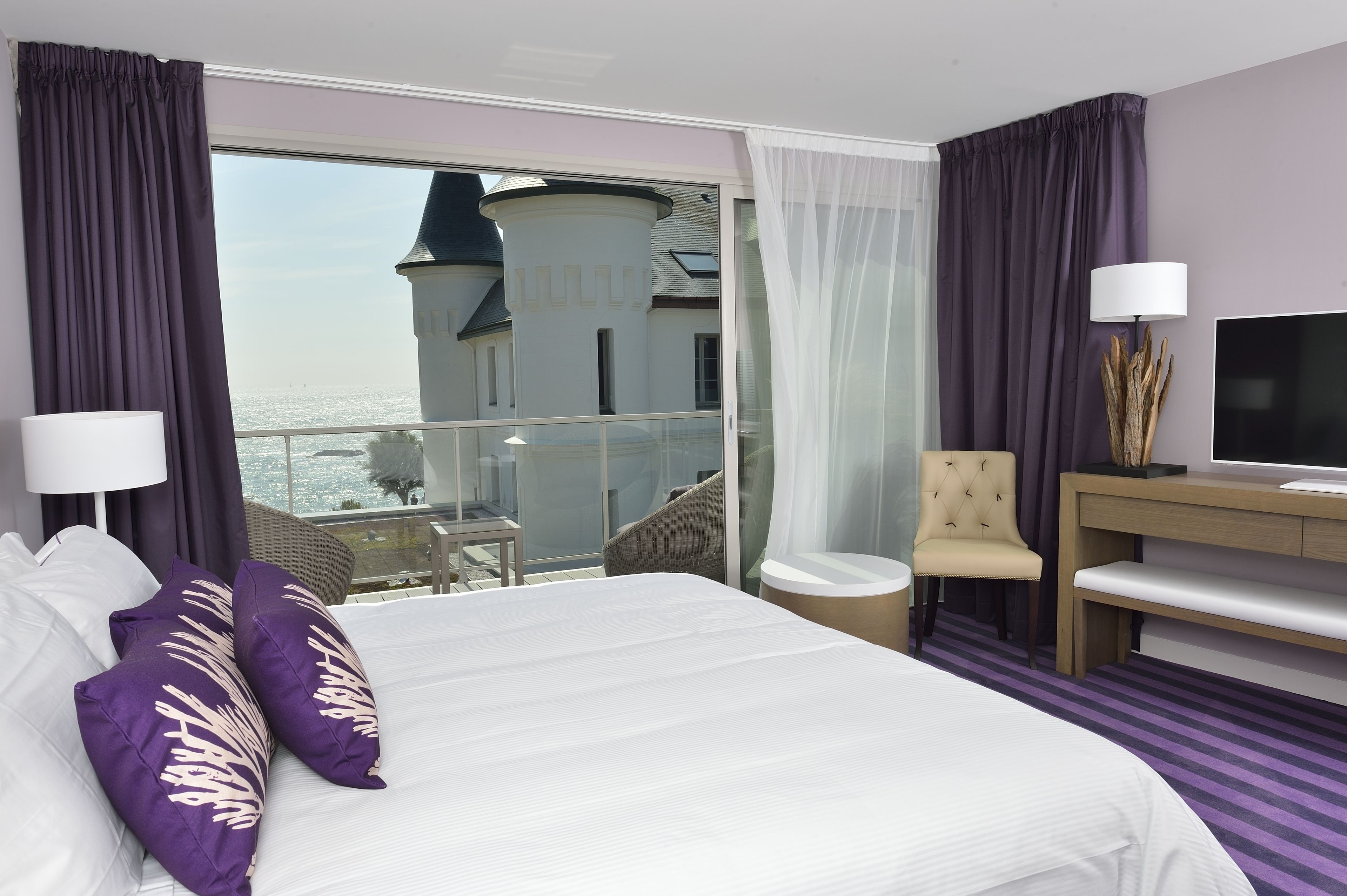 A hotel room with a large bed, a desk, a chair, and a TV. There is a balcony with a view of a building.
