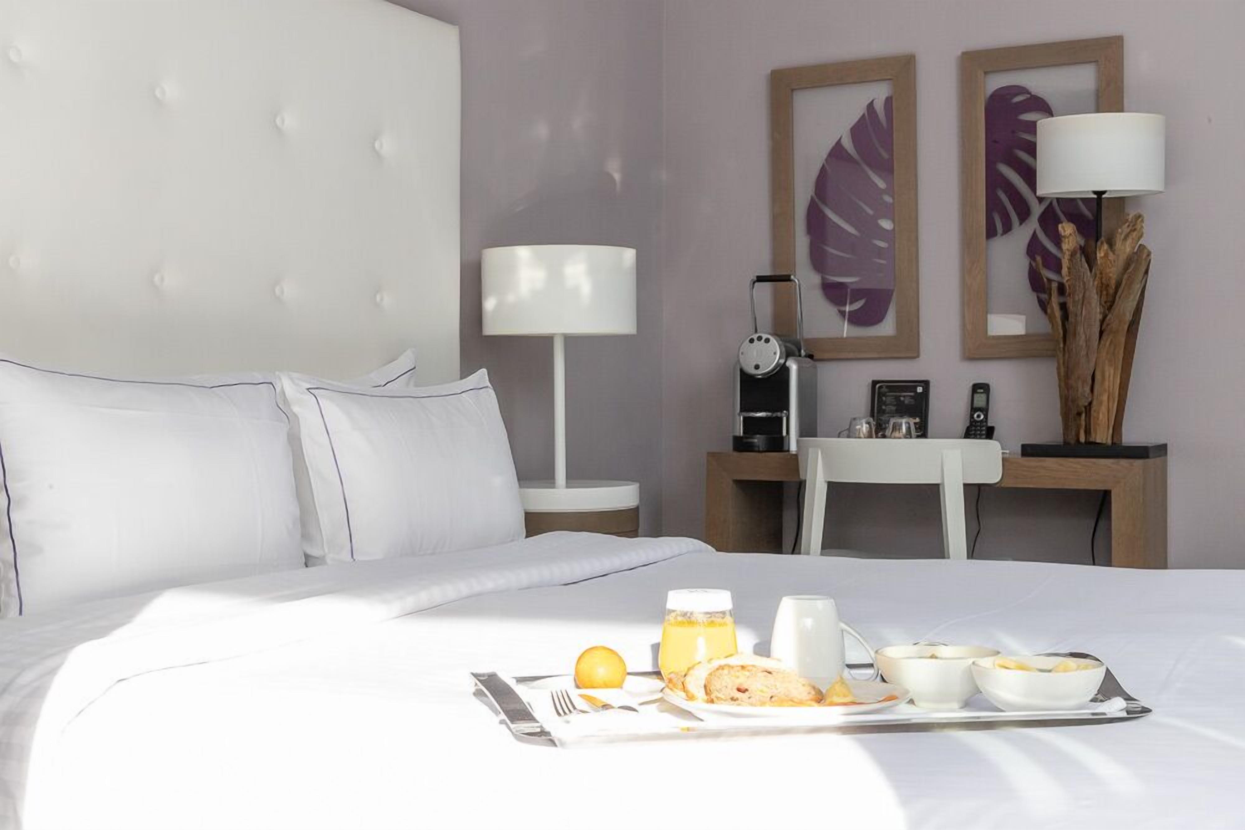 A hotel room with a bed, a bedside table with a lamp, a small table with a coffee maker, and a tray with breakfast items.
