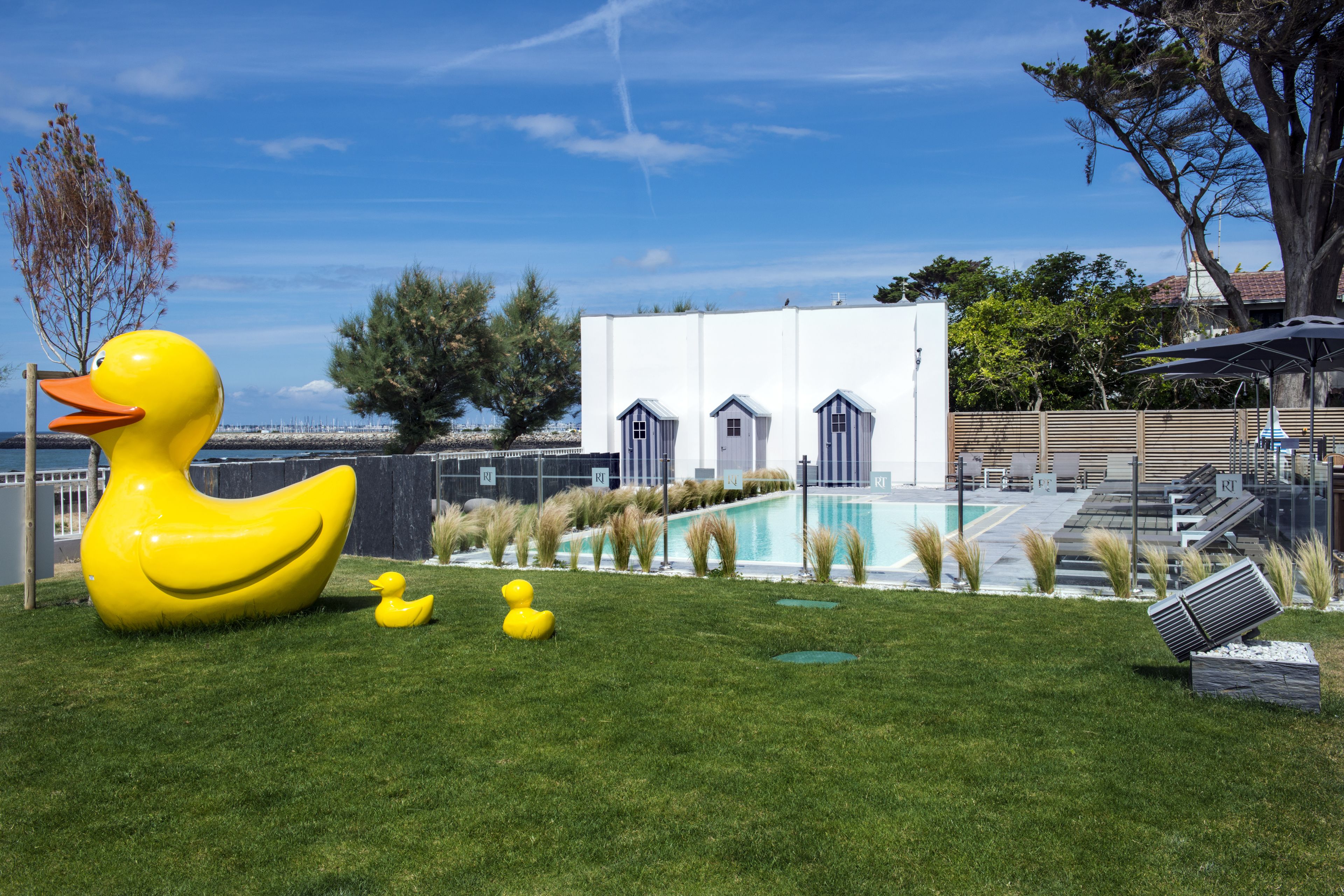 Un espace piscine agrémenté d’une grande sculpture de canard jaune, d’une petite piscine et d’un bâtiment blanc doté de nombreuses portes.