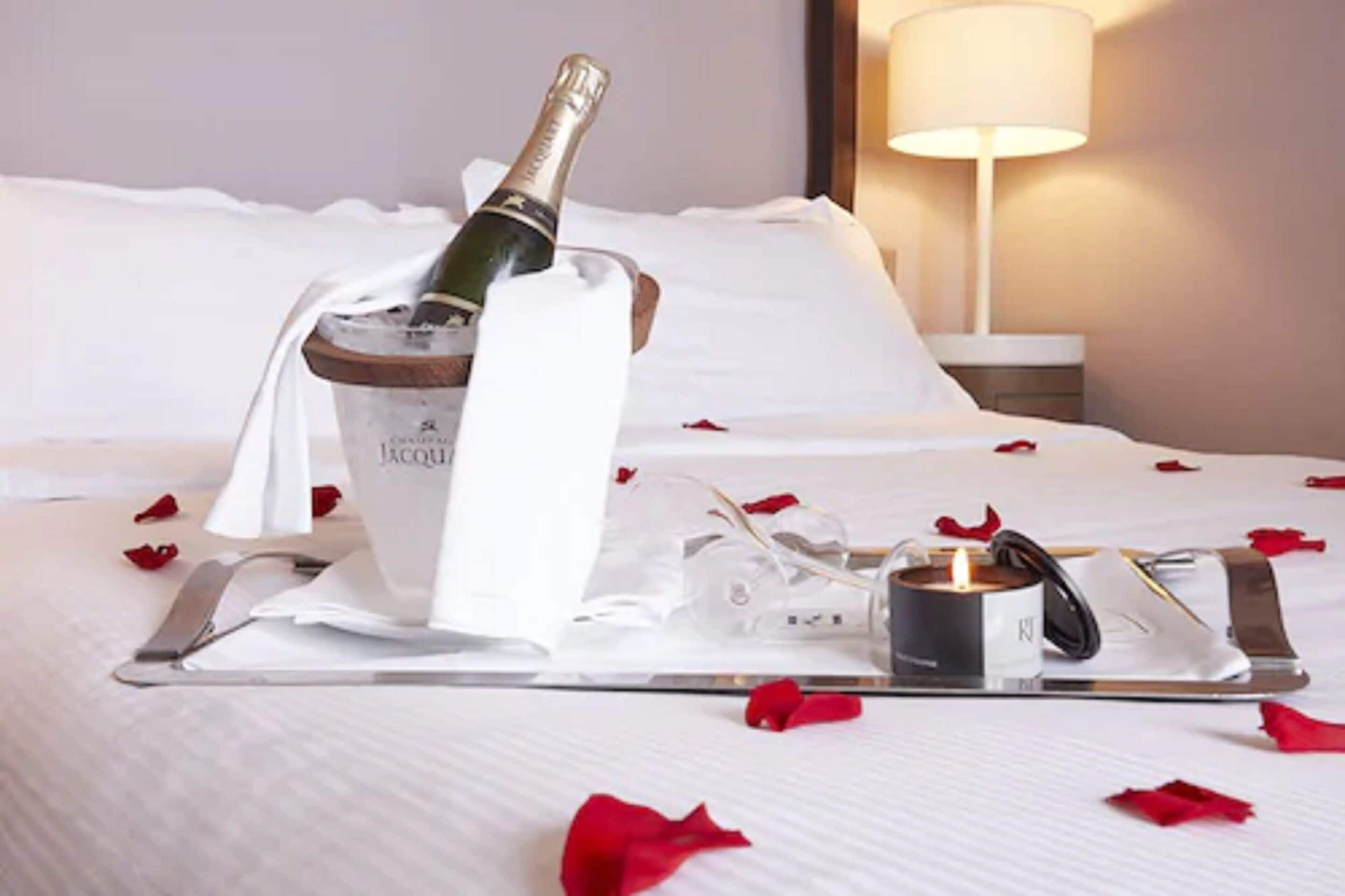A neatly made bed with a champagne bottle, glasses, and a candle on a tray, surrounded by rose petals.