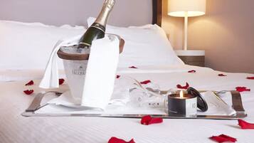 A neatly made bed with a champagne bottle, glasses, and a candle on a tray, surrounded by rose petals.