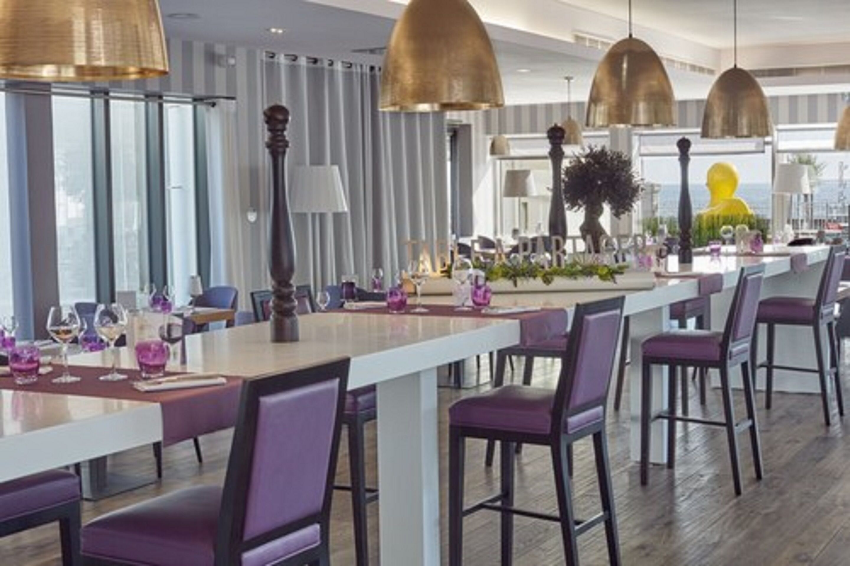 Un comedor moderno con mesas largas, sillas moradas y grandes lámparas colgantes.