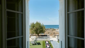 Vista desde una habitación con un ventanal grande que da a una playa, un árbol y un patio con mobiliario exterior.
