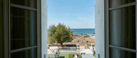 A view from a room with a large window overlooking a beach, a tree, and a patio area with outdoor furniture.