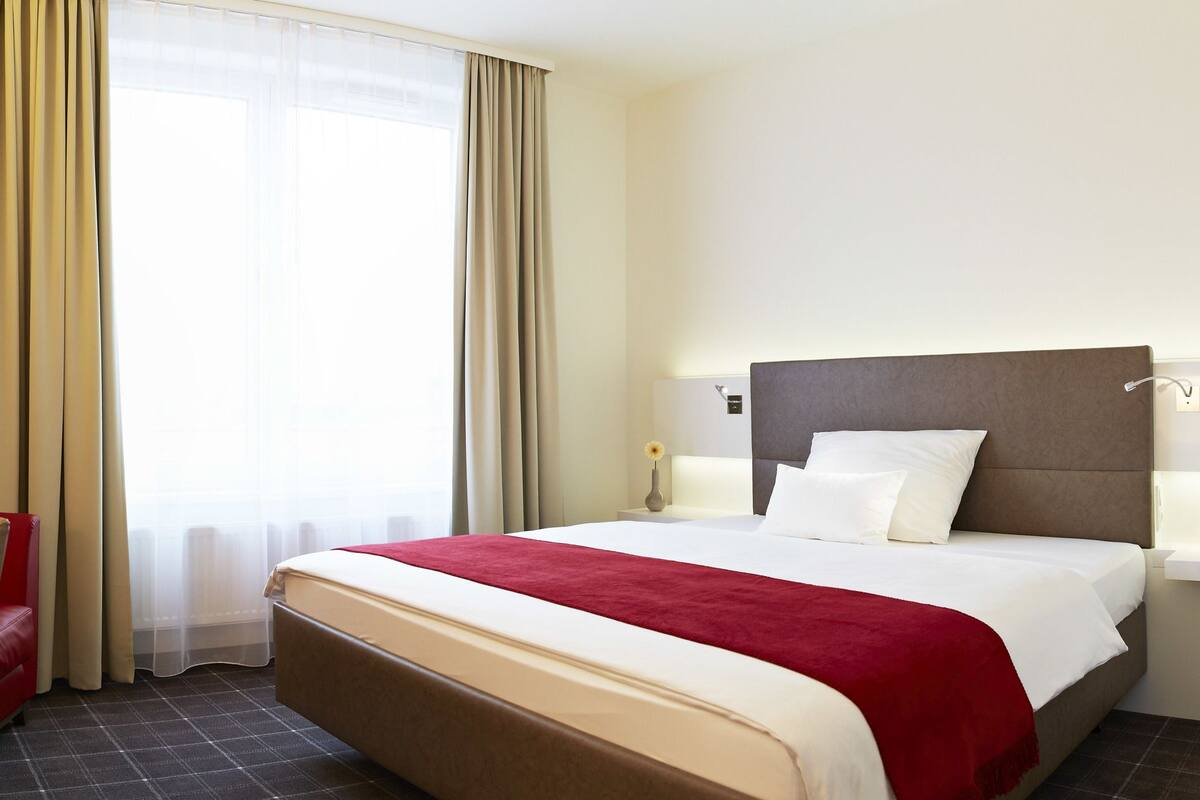 A hotel room with a bed, a desk with a lamp, a chair, and a window with curtains.