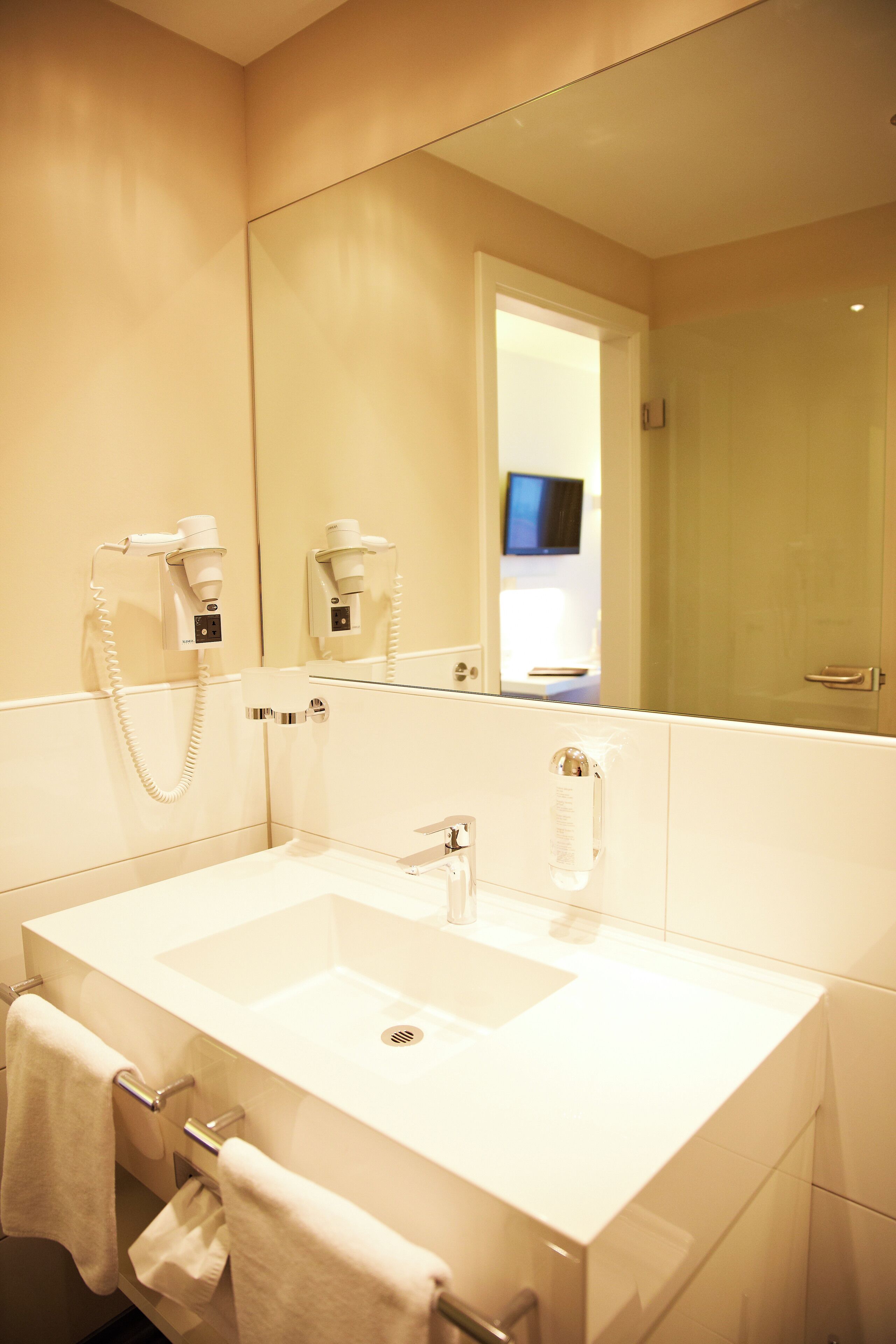 A modern bathroom with a large mirror, a white sink, and a hairdryer mounted on the wall.