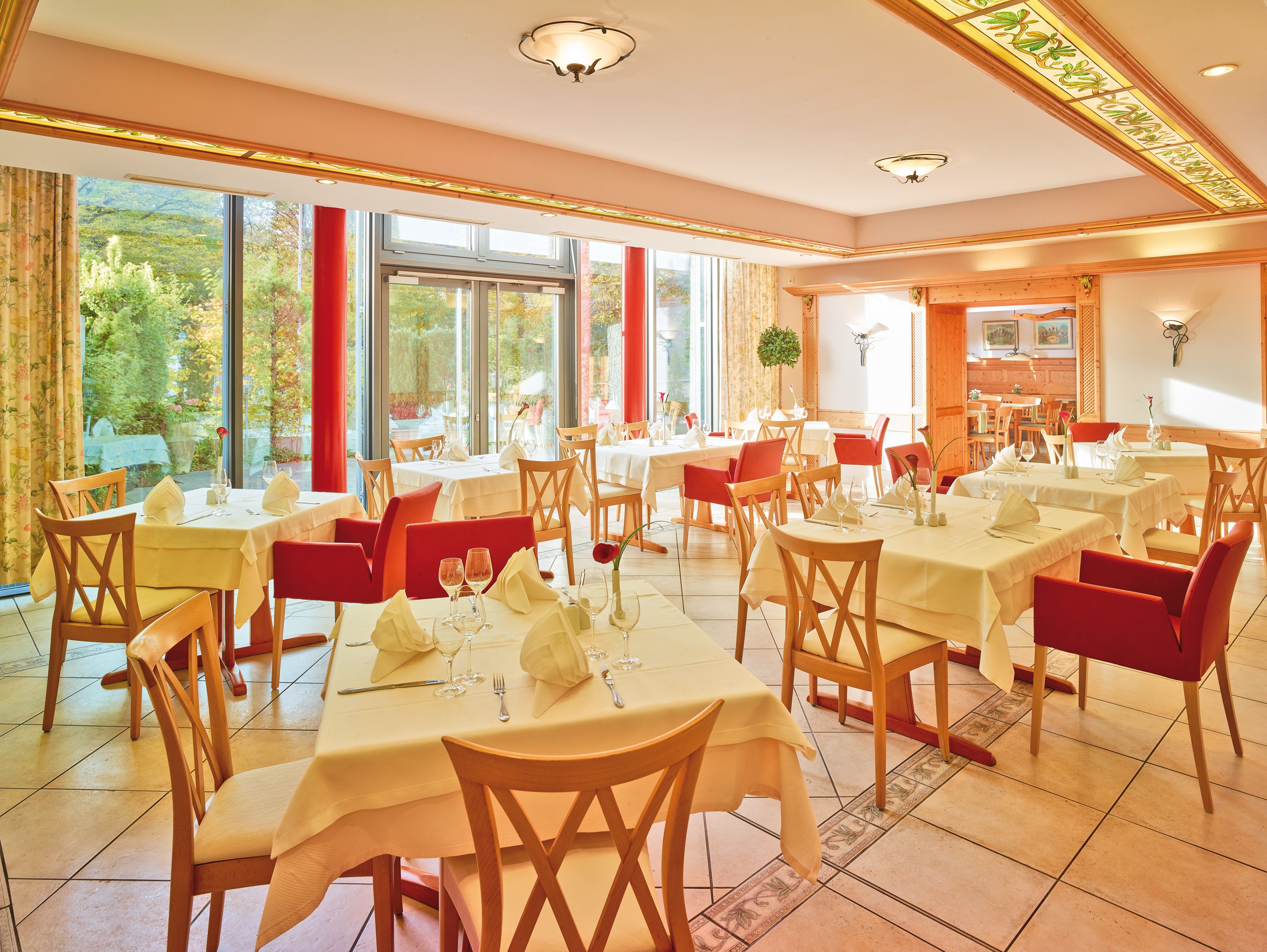 Un restaurante con varias mesas y sillas, ventanas amplias con cortinas y vistas a zonas verdes.
