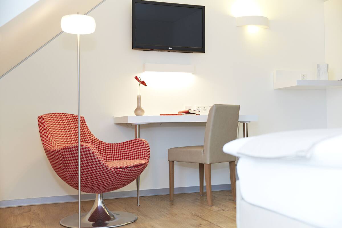 A modern room with a flat-screen TV, a red houndstooth chair, a beige chair, and a white ottoman.