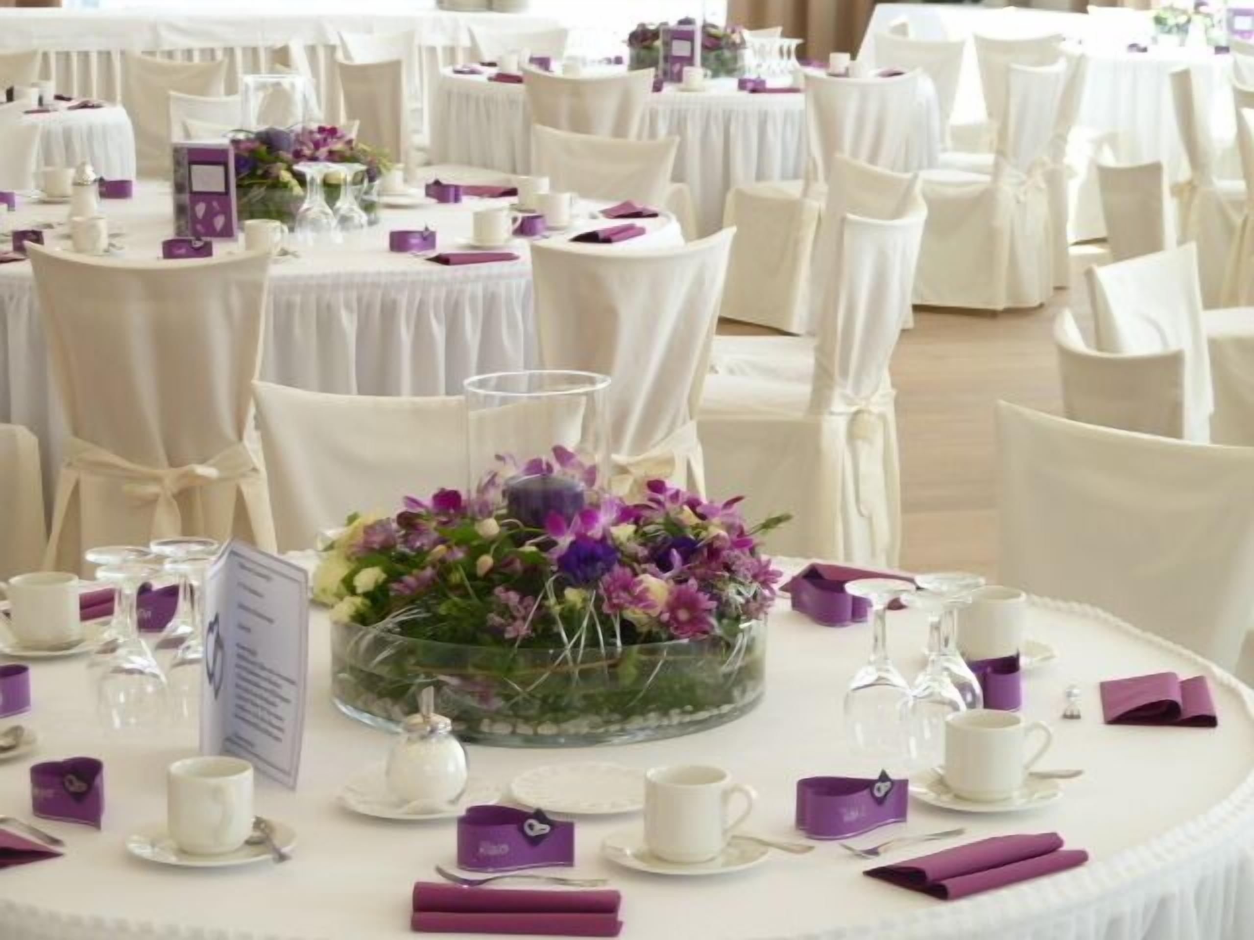 Un salón de banquetes con mesas redondas dispuestas para un evento formal, con manteles blancos, servilletas moradas y centros de mesa florales.
