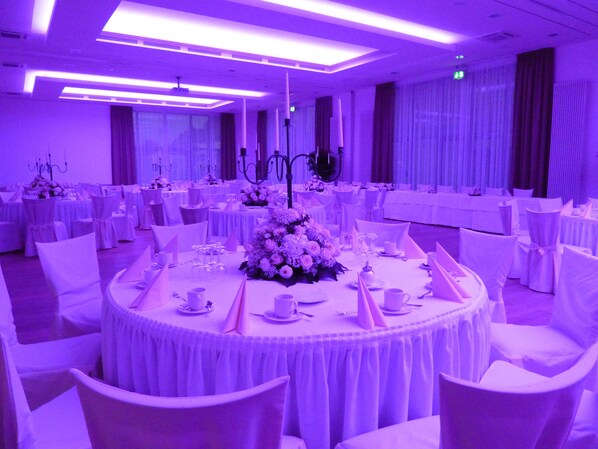 A banquet hall with round tables set for dining, white chairs, and a central floral arrangement.
