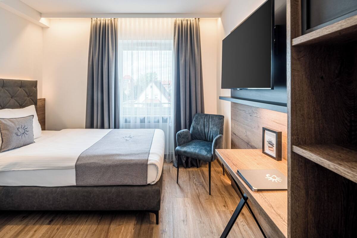 A modern hotel room with a bed, a chair, a desk with a laptop, a flat-screen TV, and a window with curtains.