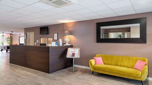 Un espace d’accueil comprenant un comptoir de réception en bois, un canapé jaune, un téléviseur fixé au mur et un panneau d’affichage.