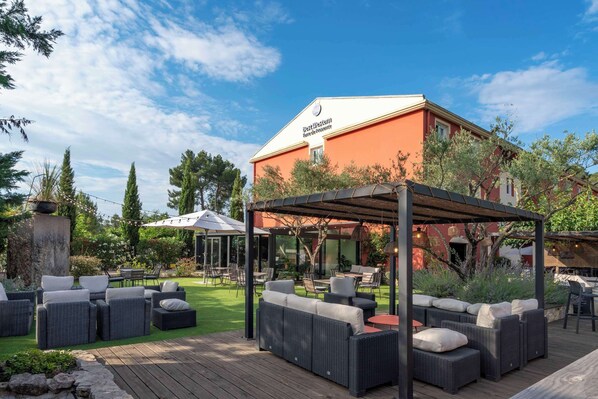 Een terras met rieten meubilair en een pergola, een gebouw met een rode gevel, en een heldere lucht.