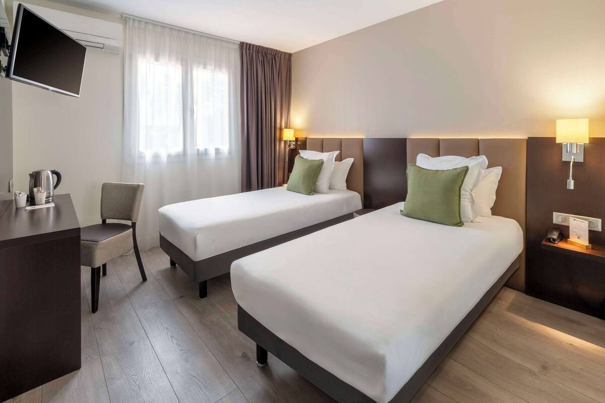 Une chambre d’hôtel avec deux lits simples, un bureau avec une chaise, un téléviseur à écran plat et une fenêtre avec des rideaux.