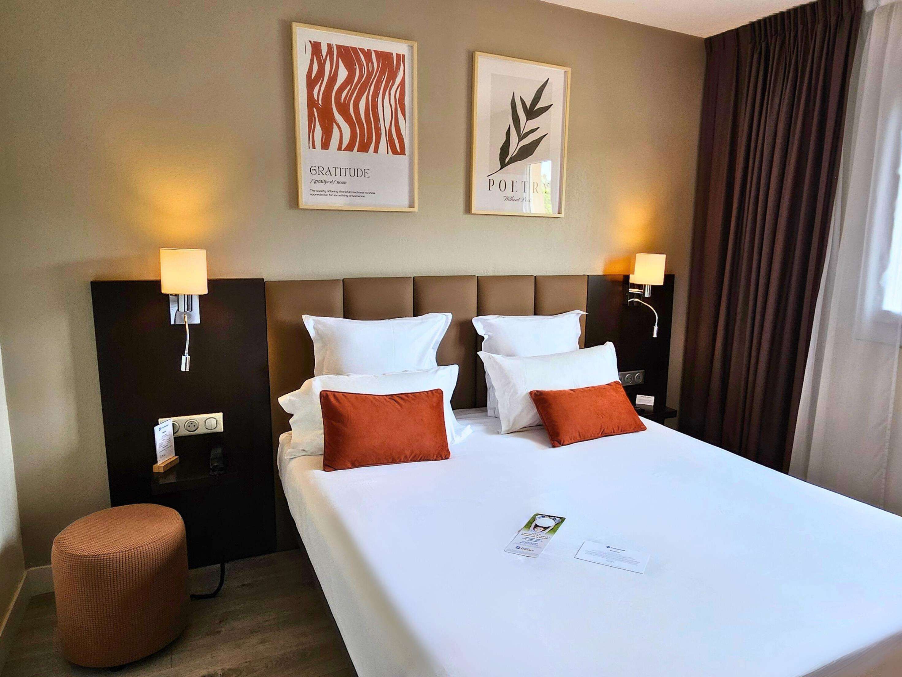 A hotel room with a large bed, two wall-mounted lamps, a nightstand, a small ottoman, and two framed artworks on the wall.