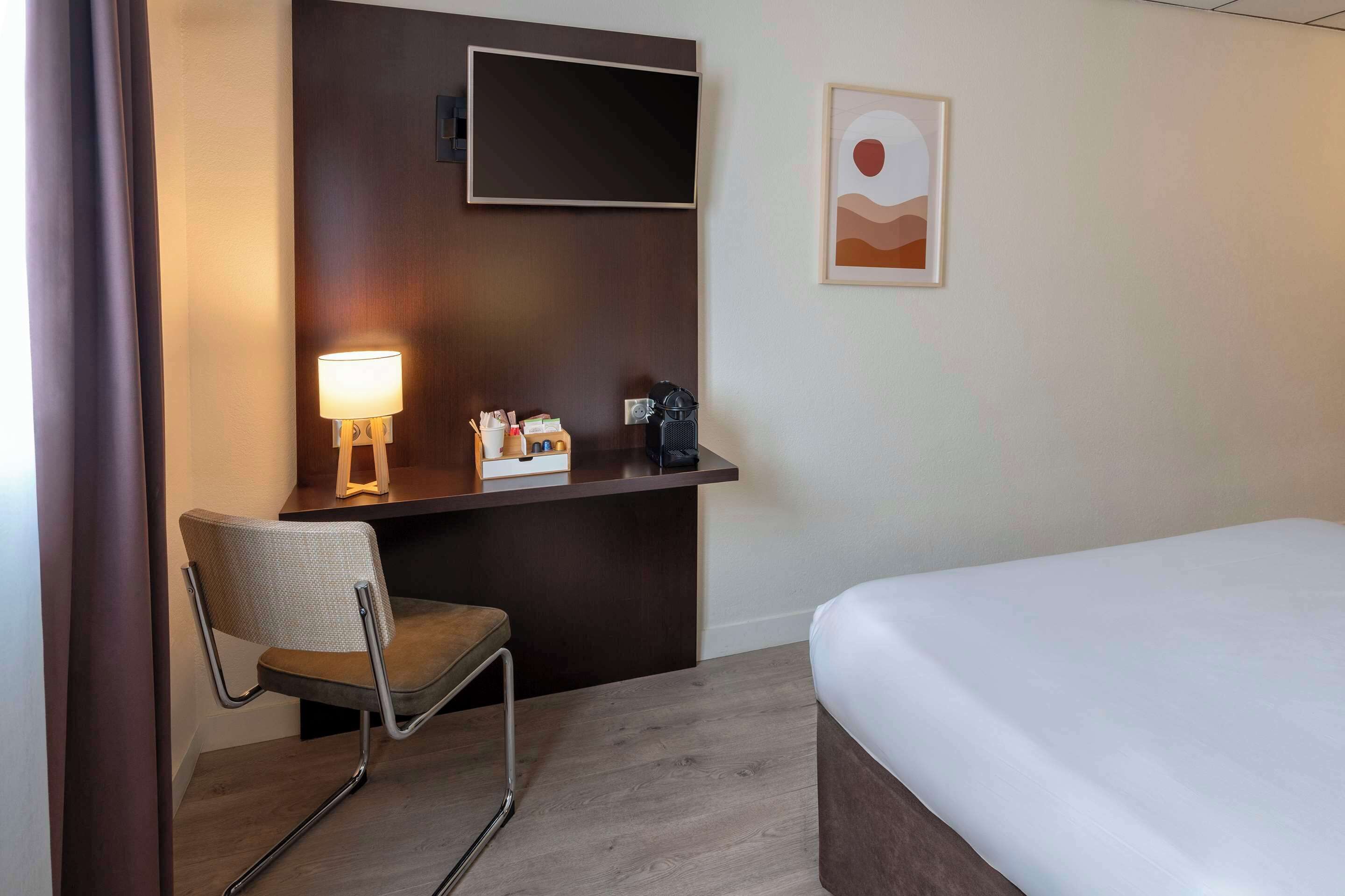 A hotel room with a bed, a desk with a lamp, a chair, a coffee machine, and a framed picture on the wall.