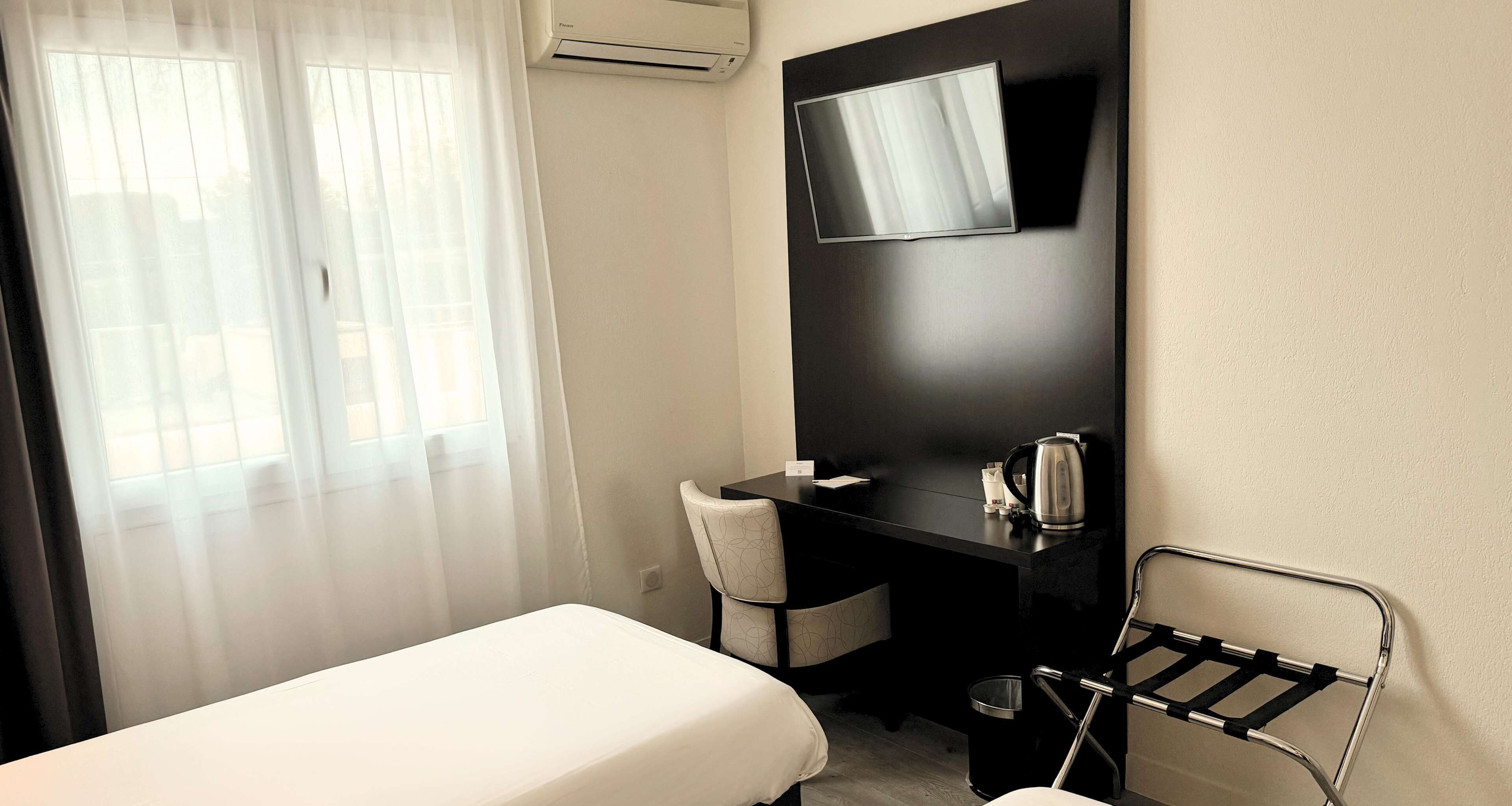 A hotel room with a bed, a desk with a chair, a TV mounted on the wall, and a window with sheer curtains.
