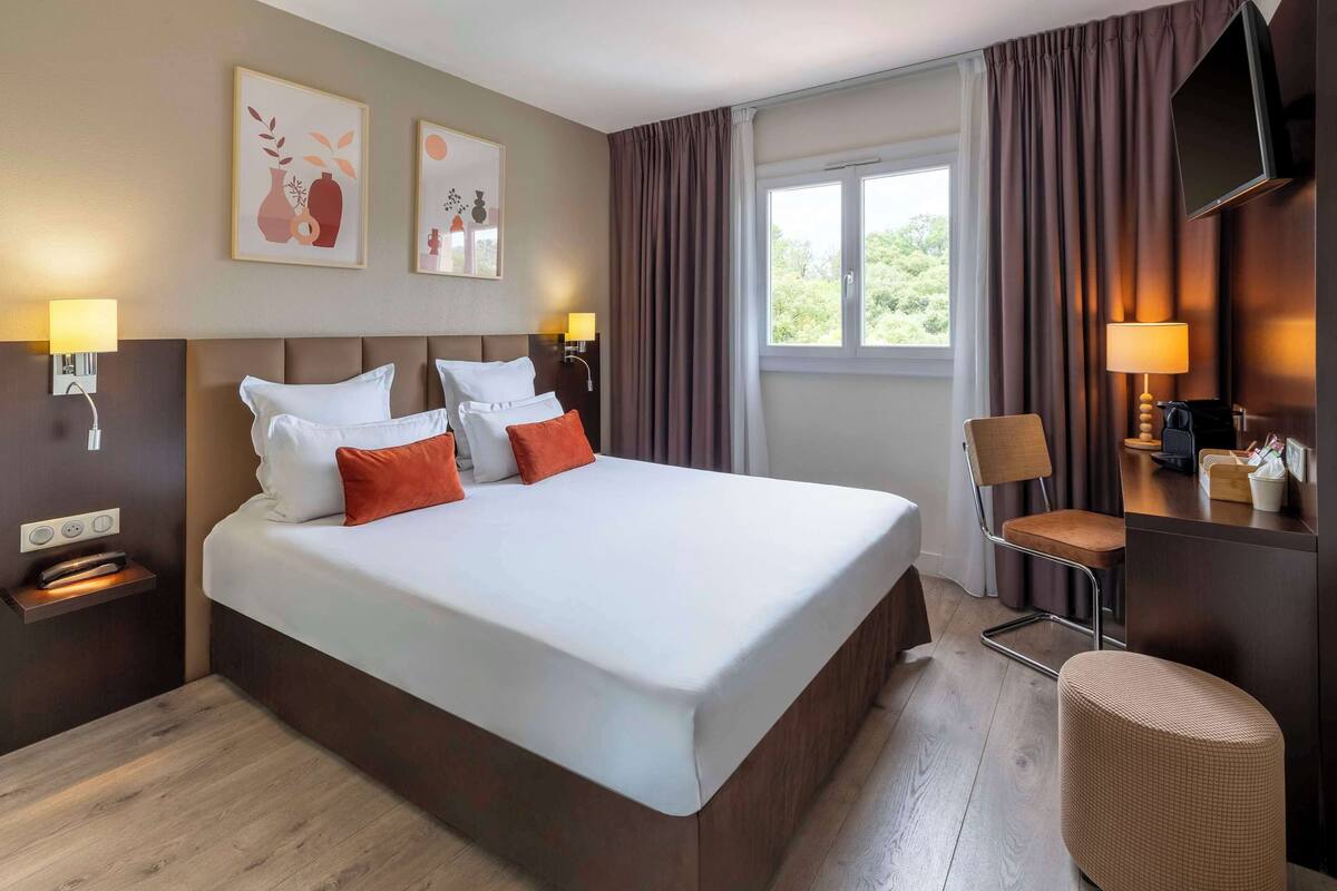 Une chambre d’hôtel avec un grand lit, deux tables de chevet, un bureau avec une chaise, une télévision et une fenêtre avec des rideaux.