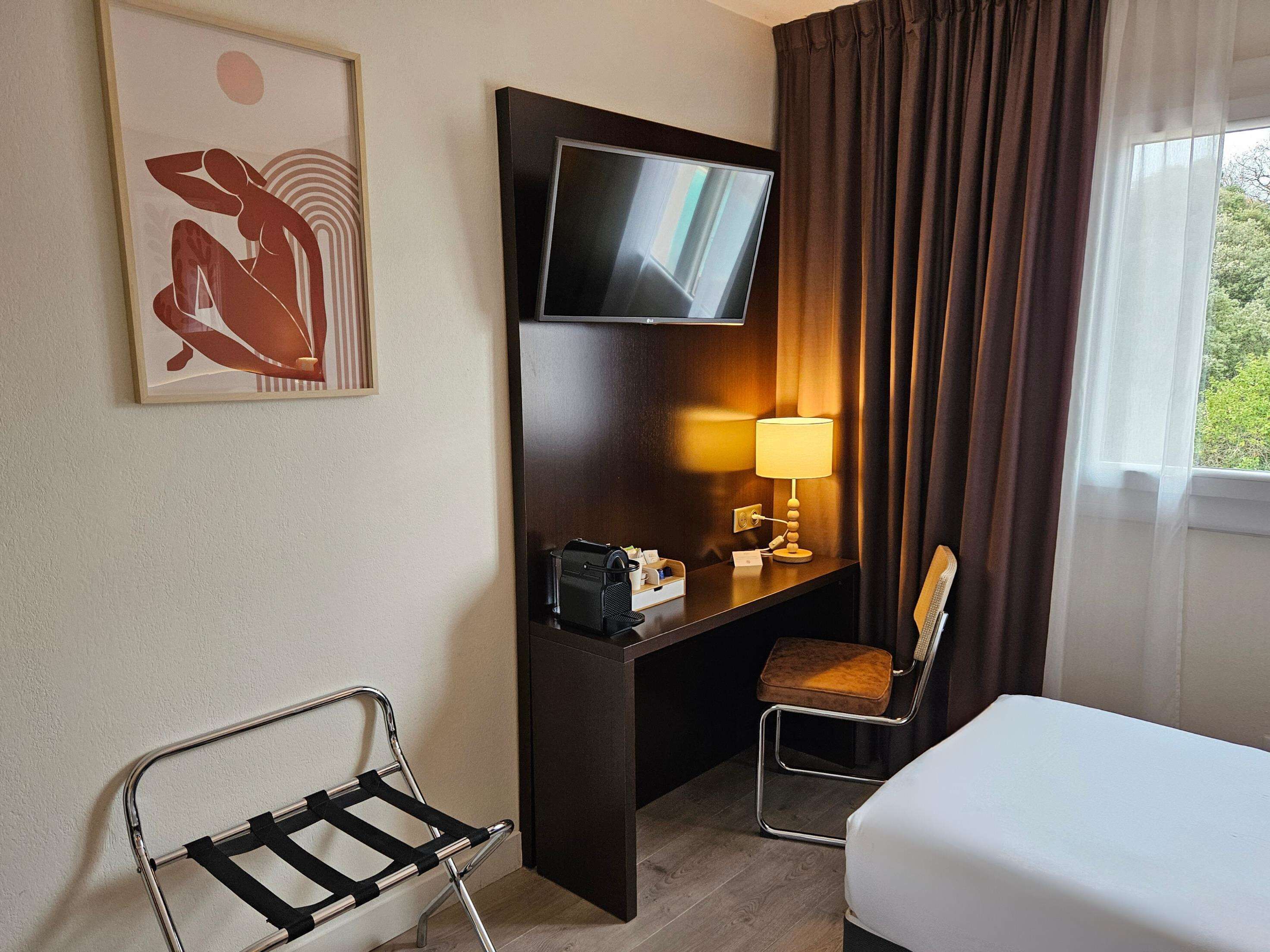 A hotel room with a bed, a desk with a TV, a chair, a lamp, and a window with curtains.
