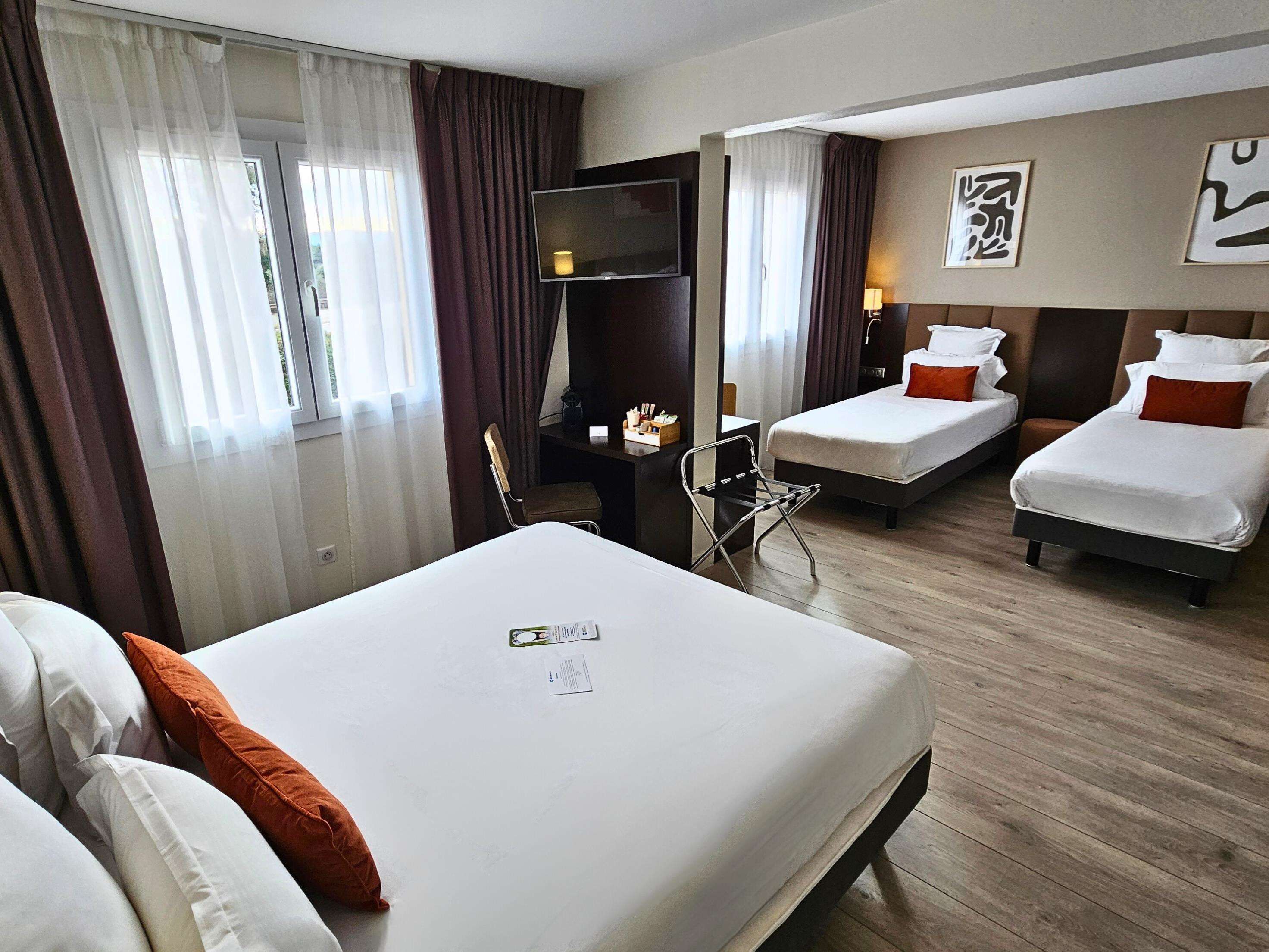 Habitación de hotel con una cama grande, dos camas más pequeñas, un escritorio, un televisor y una ventana con cortinas.