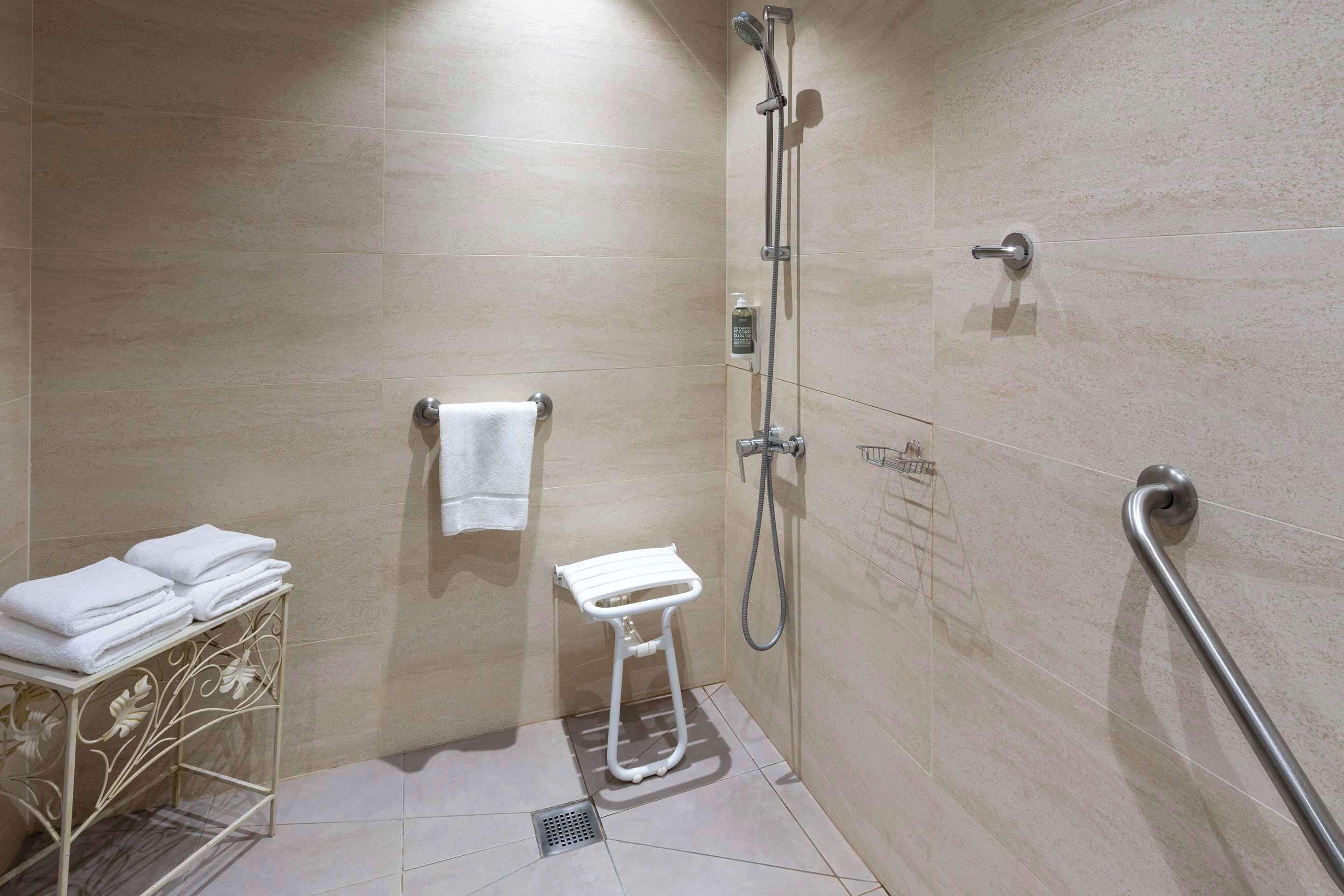 A small, modern bathroom with a shower, a toilet, and a sink. There is a towel rack with folded towels and a small table with a decorative metal frame.