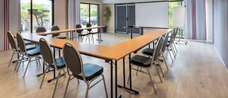 Un salón de conferencias con una larga mesa, sillas, una pantalla de proyector y ventanas grandes con cortinas.
