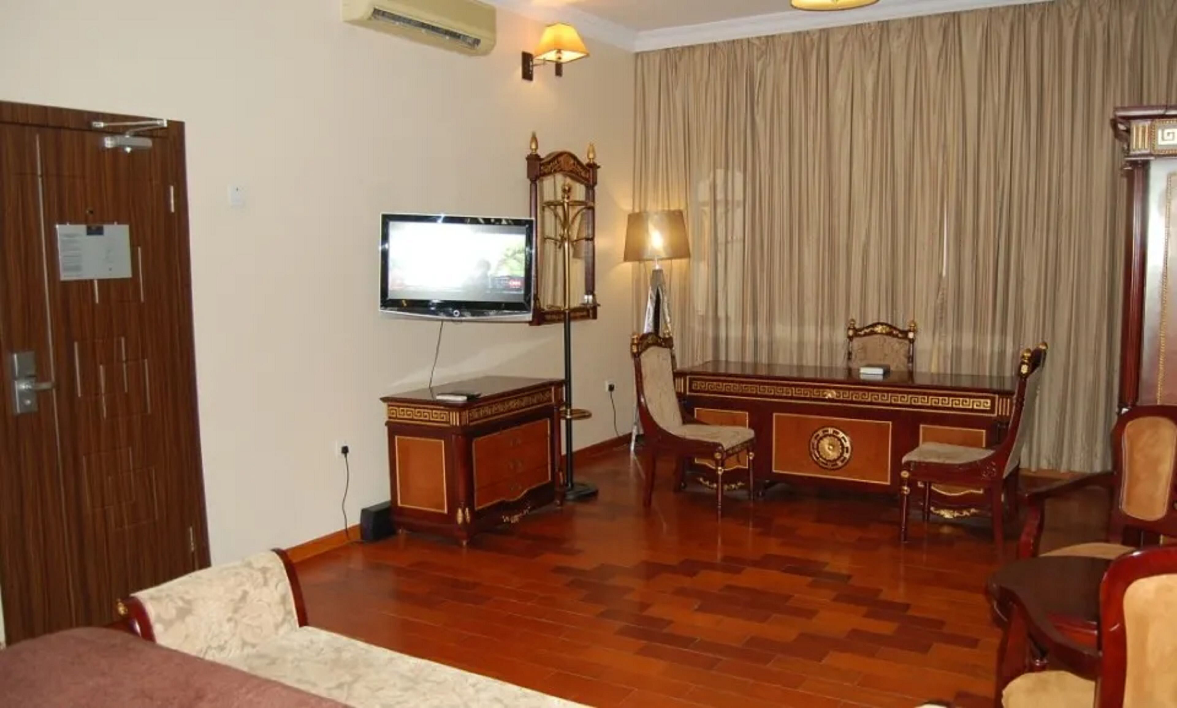 A hotel room with a wooden floor, a desk with a chair, a television, a lamp, and a bed.
