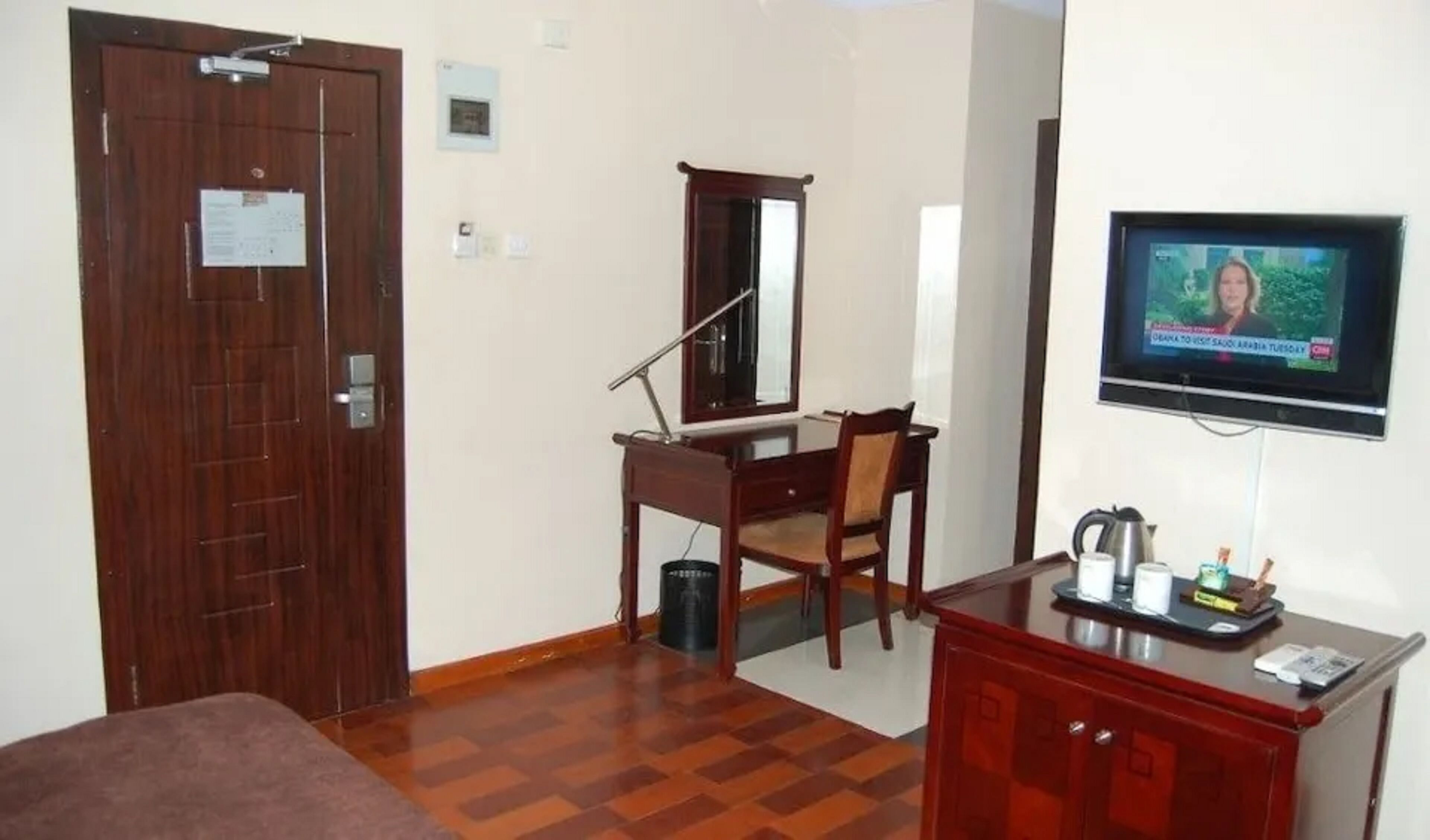 Une chambre d’hôtel avec une porte en bois, un bureau avec une chaise, un téléviseur fixé au mur et une petite armoire contenant une bouilloire.