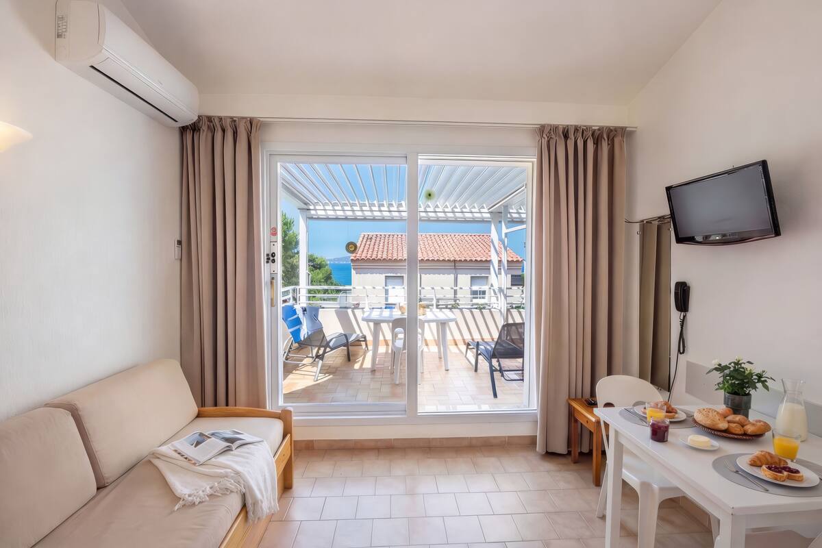 Una habitación con sofá, una mesa de desayuno, televisión y vistas a través de un ventanal grande.