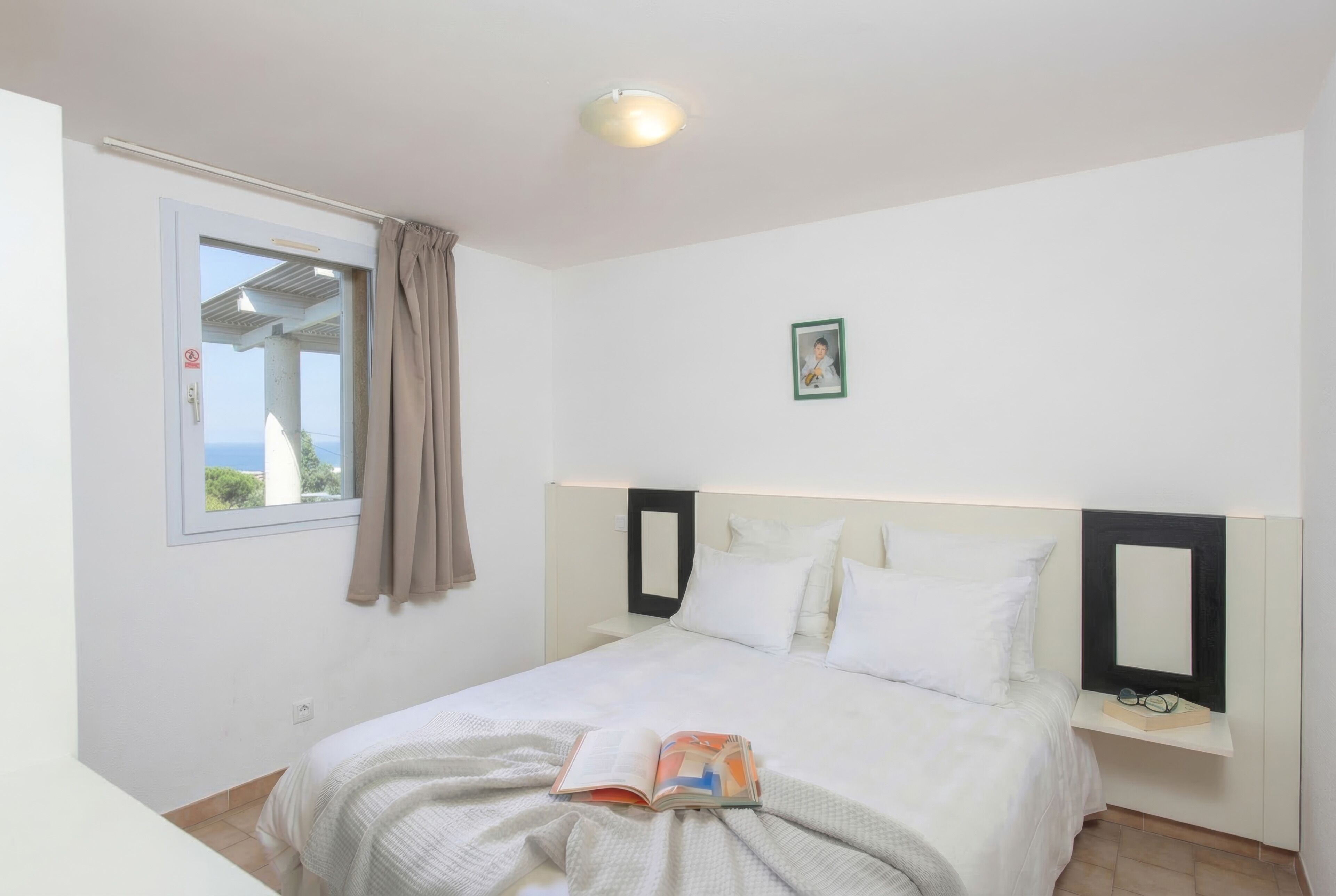 Een slaapkamer met een bed, een raam met gordijnen, een nachtkastje met een spiegel en een lijst met een foto aan de muur.