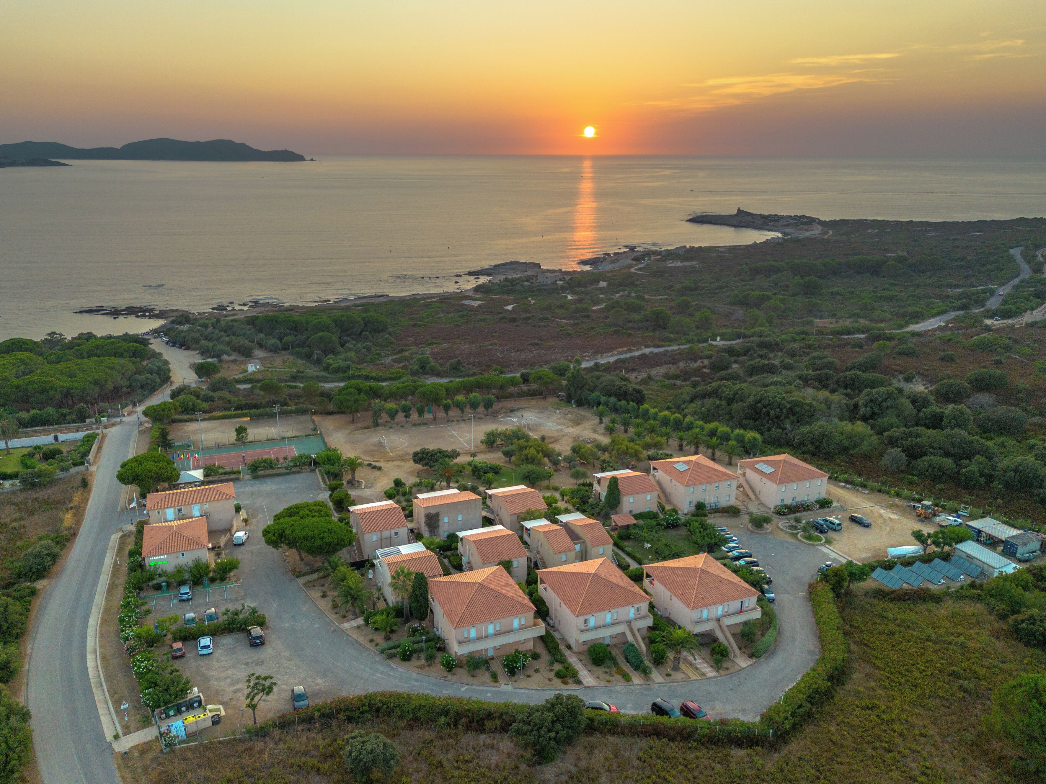 A coastal residential area with houses, a road, and a sunset over the sea.