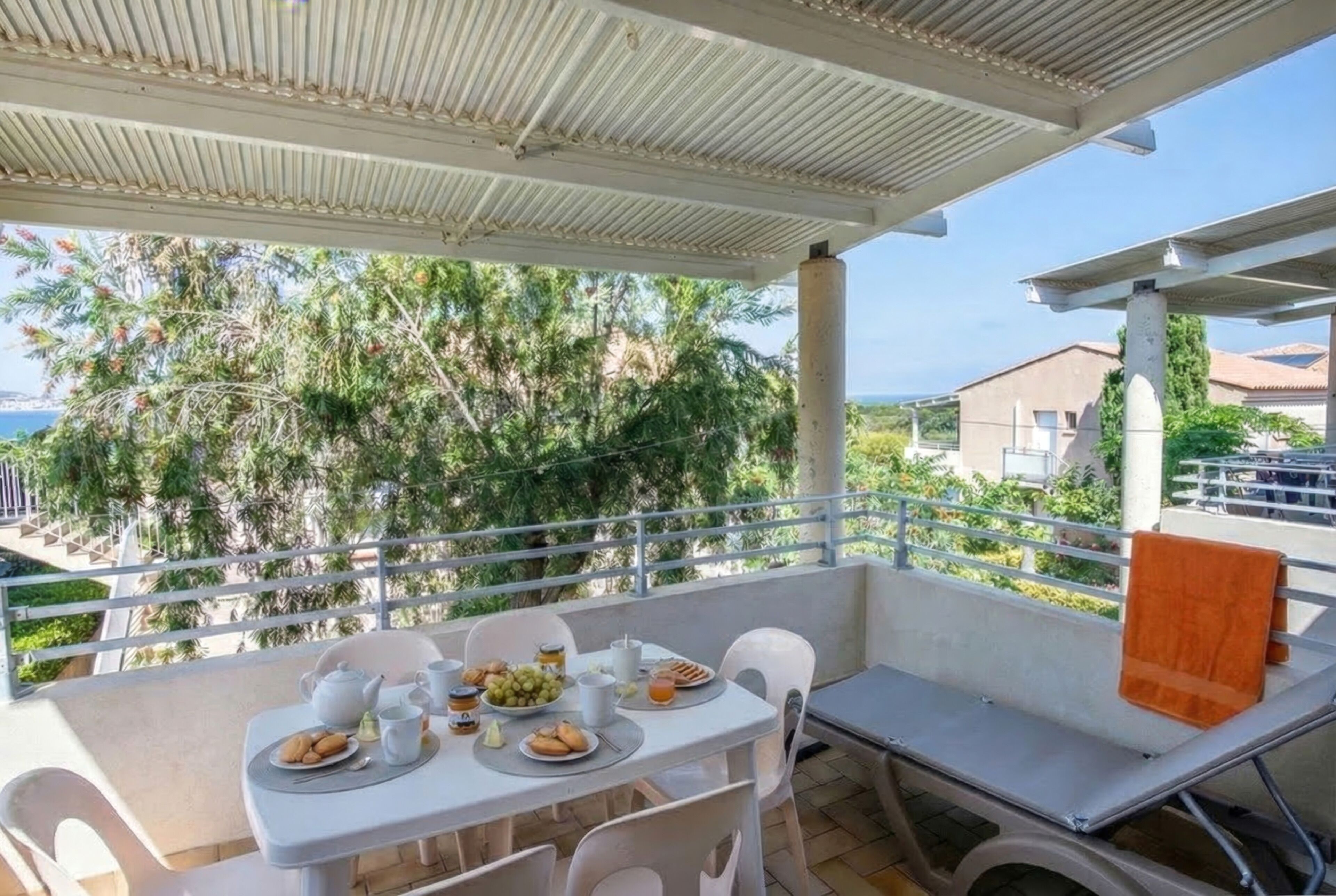 A covered outdoor dining area with a table set for two, chairs, and a view of greenery and buildings in the background.
