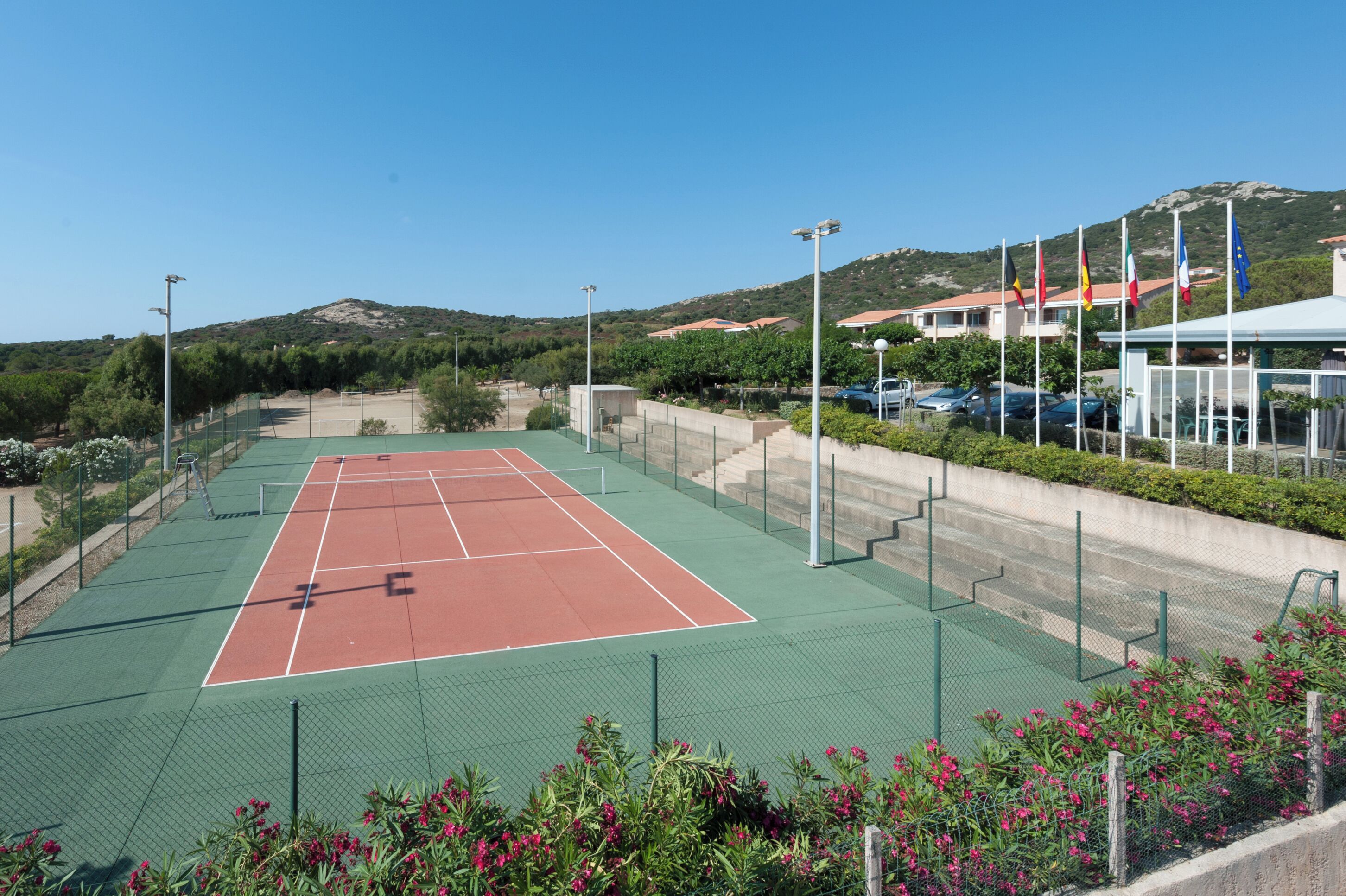 A tennis court with a green and red surface, surrounded by a fence and a row of flags.