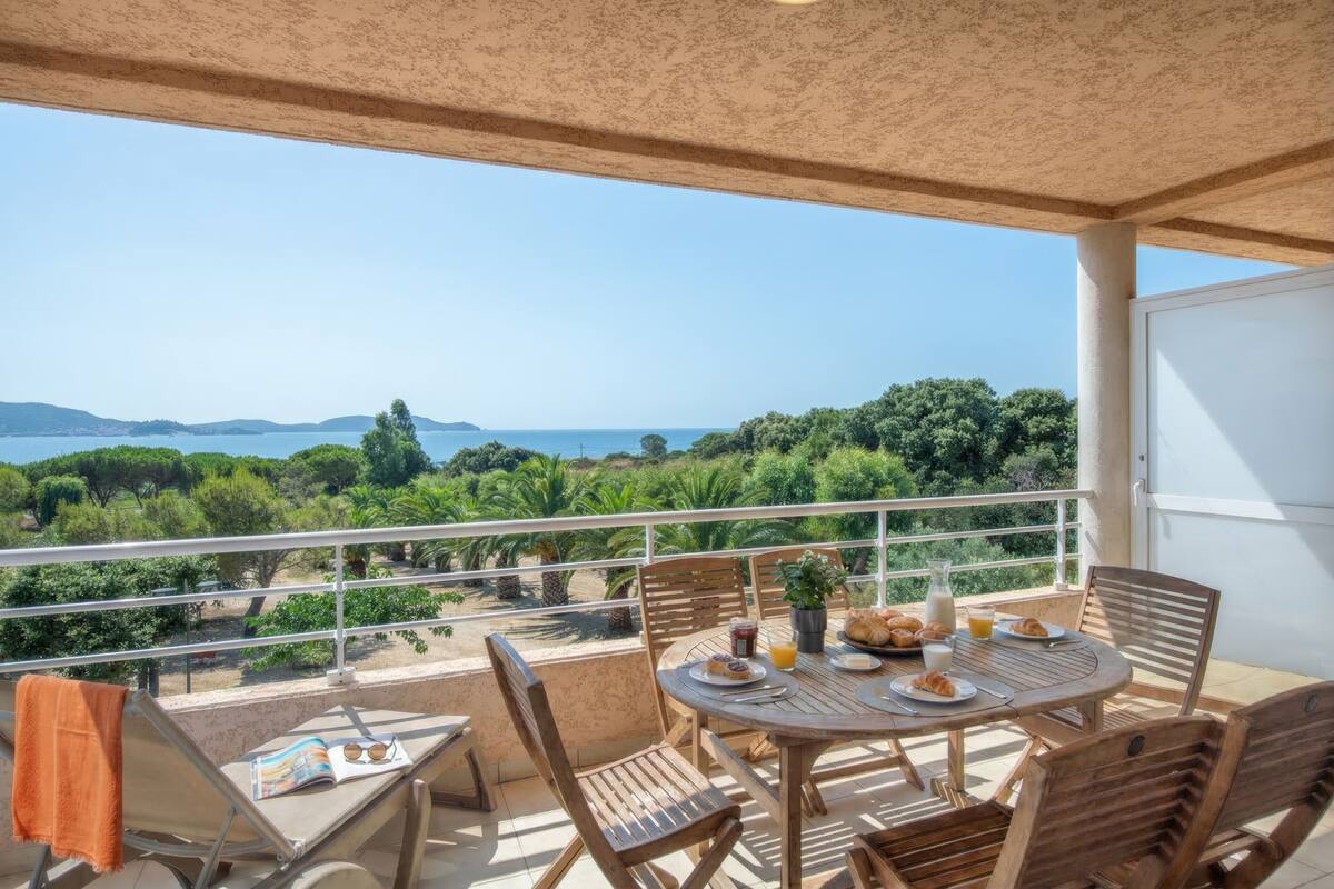Un balcón con una mesa lista para el desayuno, una silla con una toalla y vistas al mar y la vegetación.