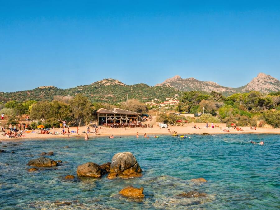 A coastal beach with clear waters, rocky outcrops, and a building with outdoor seating.