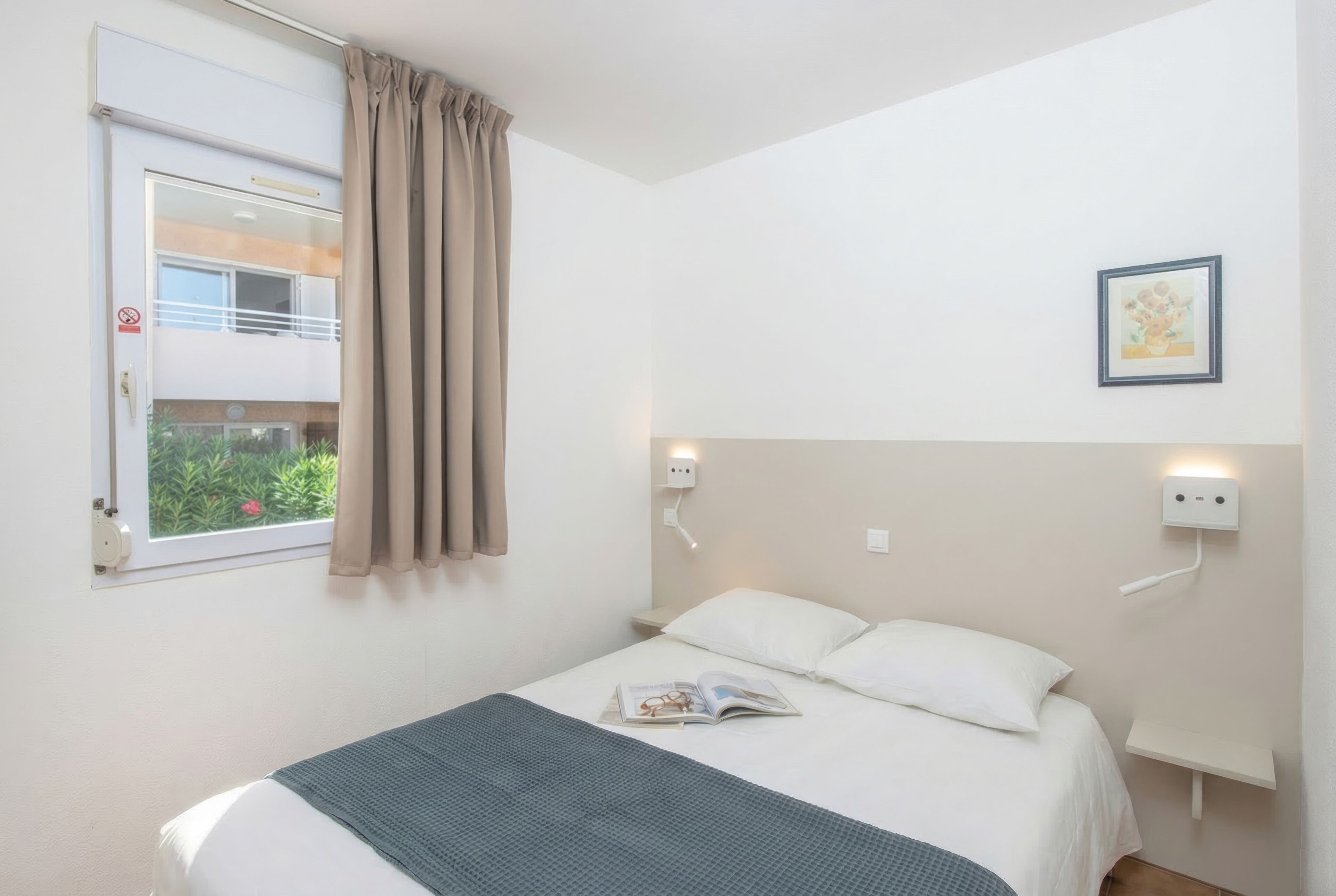 Un lit simple avec une tête de lit blanche, une fenêtre donnant sur un paysage verdoyant et un luminaire fixé au mur.