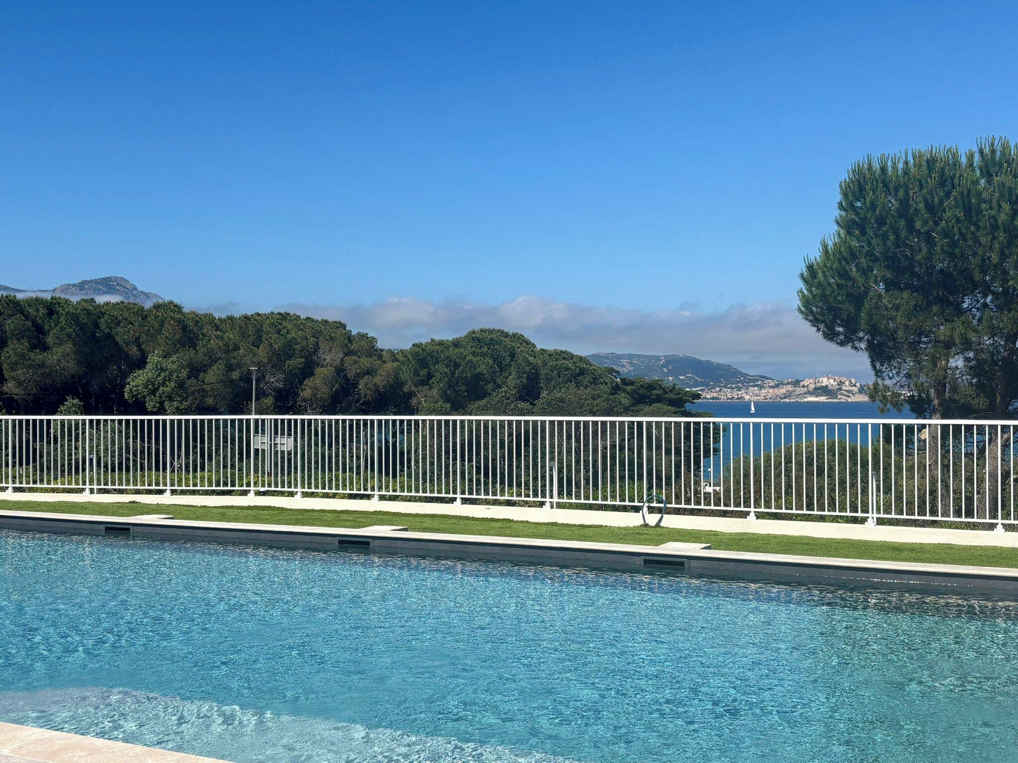 A swimming pool with a white fence, a clear blue sky, and a view of a coastal town in the background.