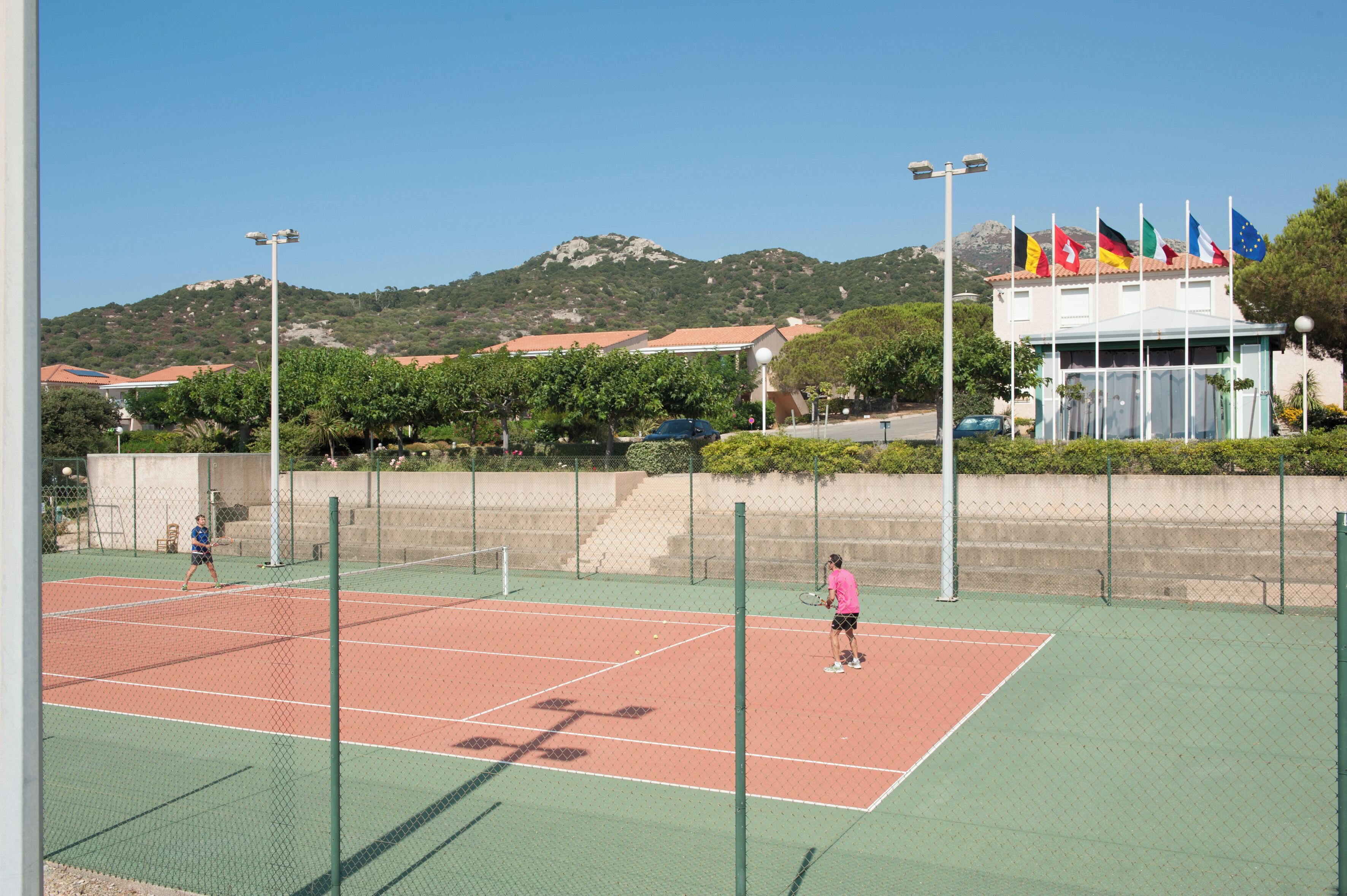 Un court de tennis en plein air, doté d’un filet et entouré d’une clôture, avec un bâtiment arborant plusieurs drapeaux en arrière-plan.
