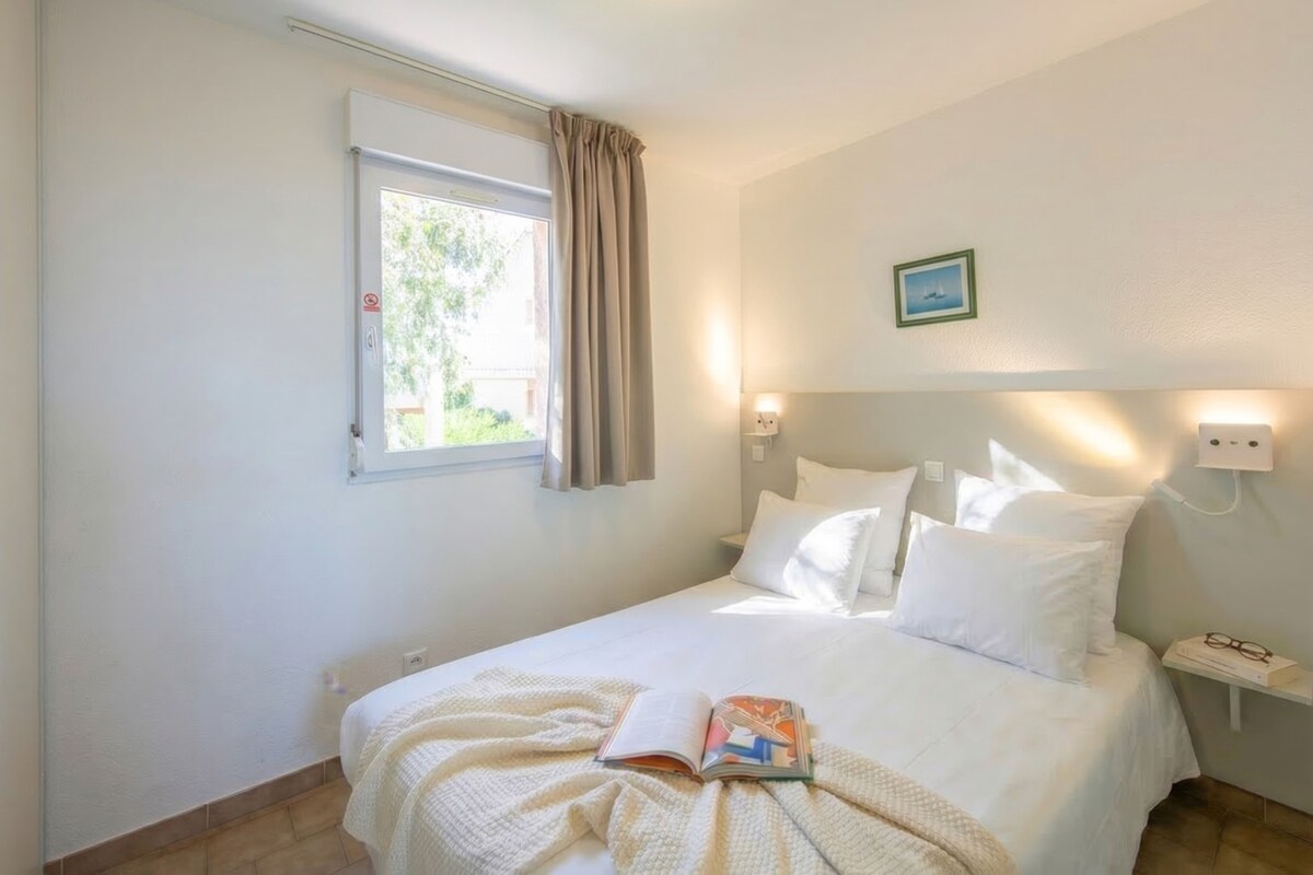 A bedroom with a bed, a window with curtains, a bedside table with a lamp, and a wall-mounted picture.