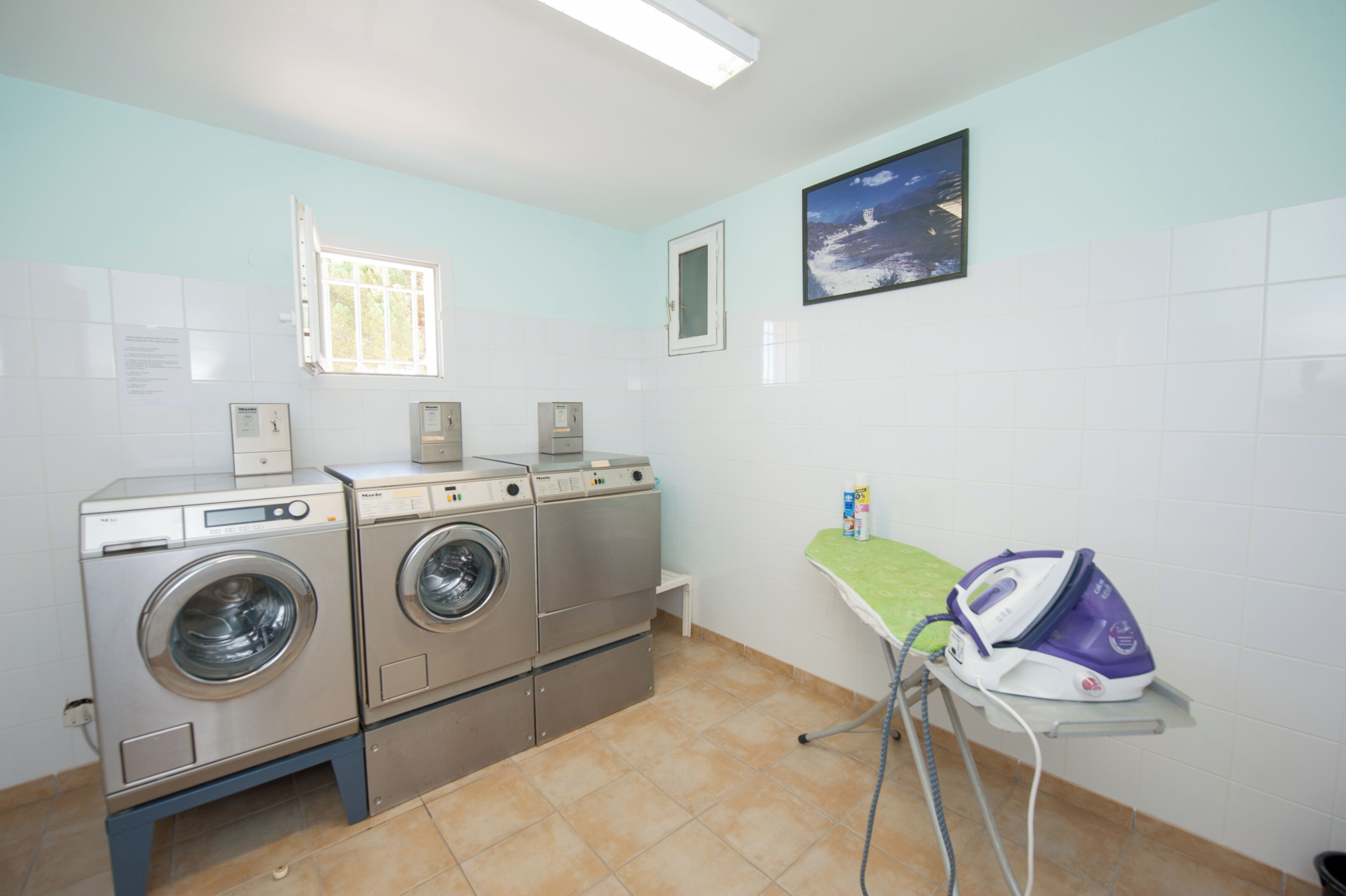 A laundry room with two front-loading washers, a small ironing station, a window, and a picture on the wall.