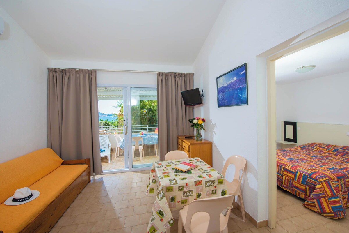 A hotel room with a sofa, a dining table with a floral tablecloth, a TV, a bed with a colorful bedspread, and a balcony with a view.