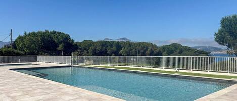 A rectangular swimming pool with a white fence, surrounded by a tiled deck, with a clear view of a mountainous landscape and a body of water in the distance.