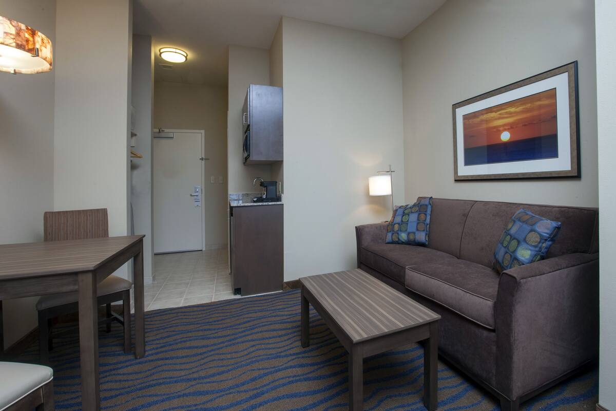 Habitación de hotel con mesa de comedor, sofá, una pequeña mesa de centro, zona de cocina y un cuadro en la pared.