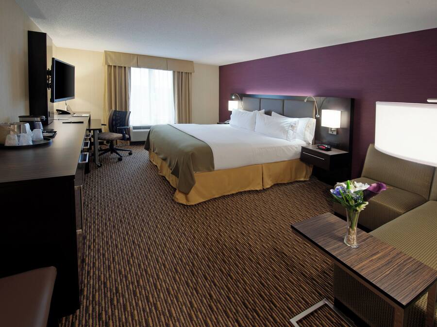 Une chambre d’hôtel avec un grand lit, un bureau avec un ordinateur, une chaise, une télévision et une petite table sur laquelle est posé un vase de fleurs.