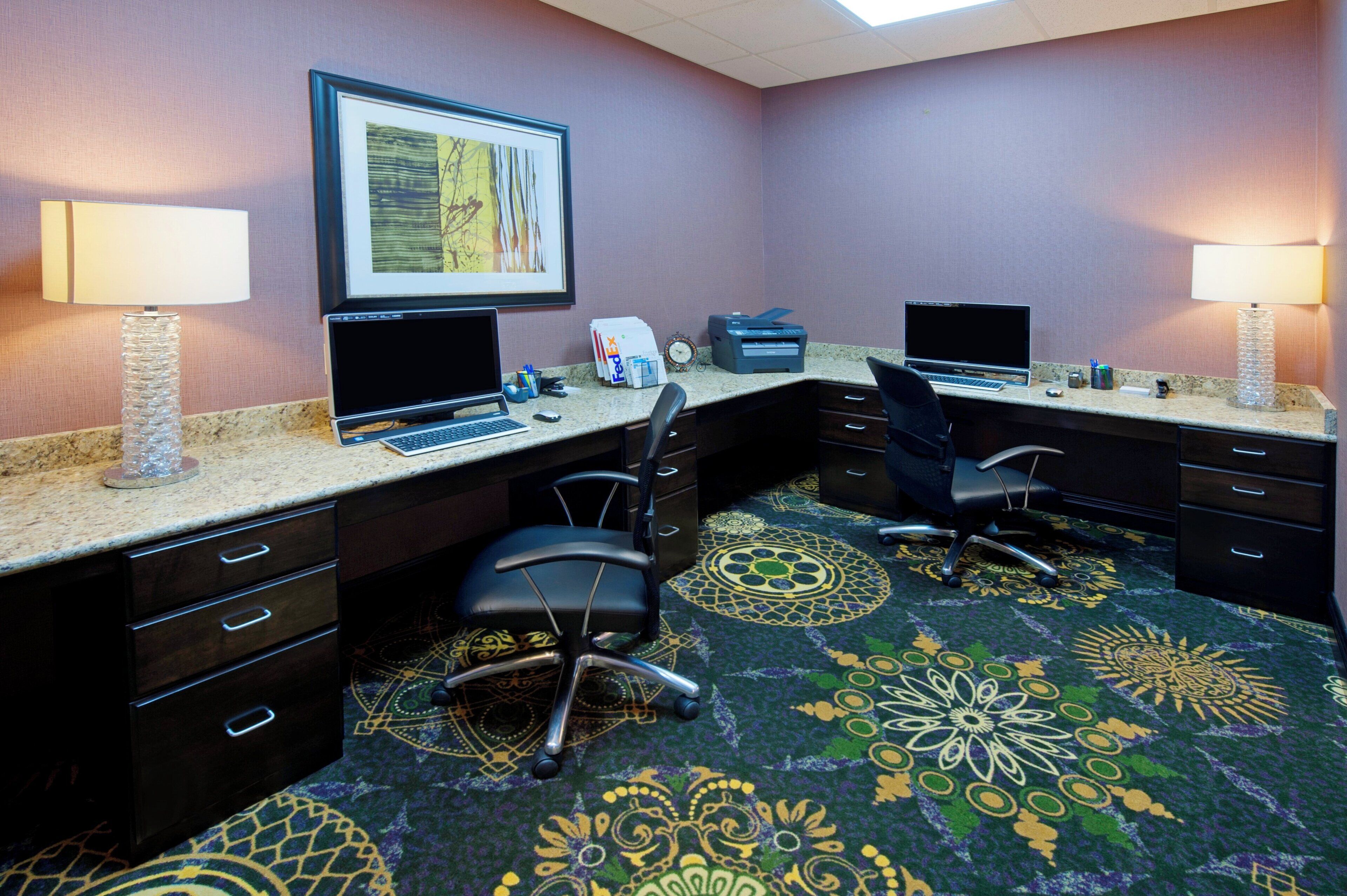 A hotel room with two desks, each with a laptop, a printer, and a lamp. There are two chairs, a carpet with a floral pattern, and a framed picture on the wall.