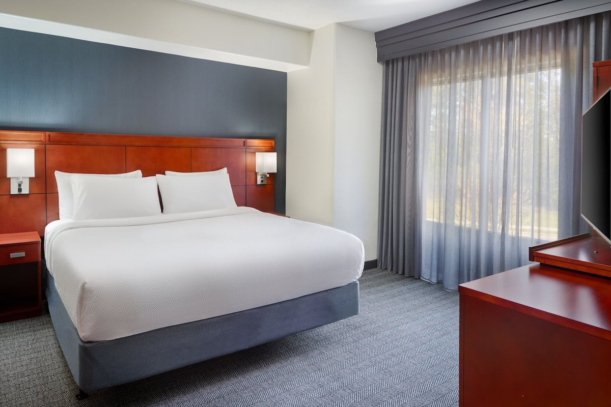Quarto de hotel com uma cama grande, cabeceira de madeira, duas mesas de cabeceira com luminárias, uma escrivaninha e uma janela ampla com cortinas.